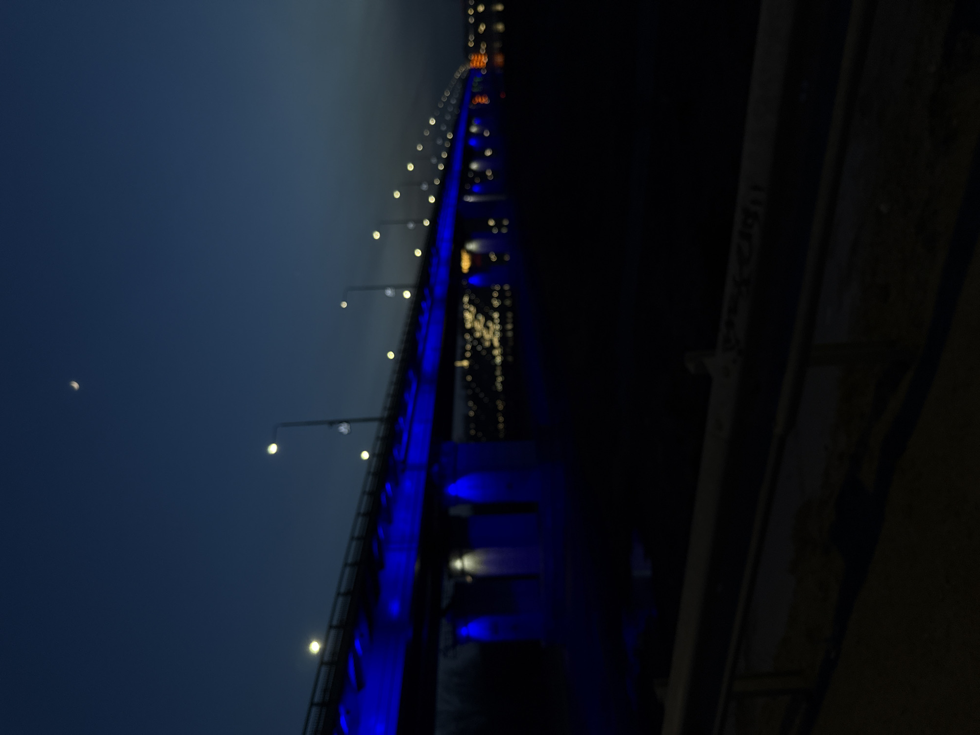 The image displays a long bridge or elevated structure at night, viewed from a slightly upward and diagonal perspective. The structure extends into the distance, characterized by a series of small, round, out-of-focus yellow-white lights lining its path. Distinct sections of the bridge are dramatically illuminated with a vibrant, electric blue light, contrasting sharply with the dark surroundings.
The sky above is a deep, dark blue, indicating either night or deep twilight. A clear crescent moon is visible in the upper left portion of the sky. To the right of the main structure, a darker, indistinct structural element, possibly a support beam or adjacent part of the bridge, is partially visible against the dark backdrop. This element has faint, unreadable markings.
No people or discernible activity are present in the static scene. The lighting suggests decorative illumination on the bridge, creating a serene and somewhat dramatic atmosphere. The weather appears clear, allowing the moon to be visible. There is no clearly legible text in the image.