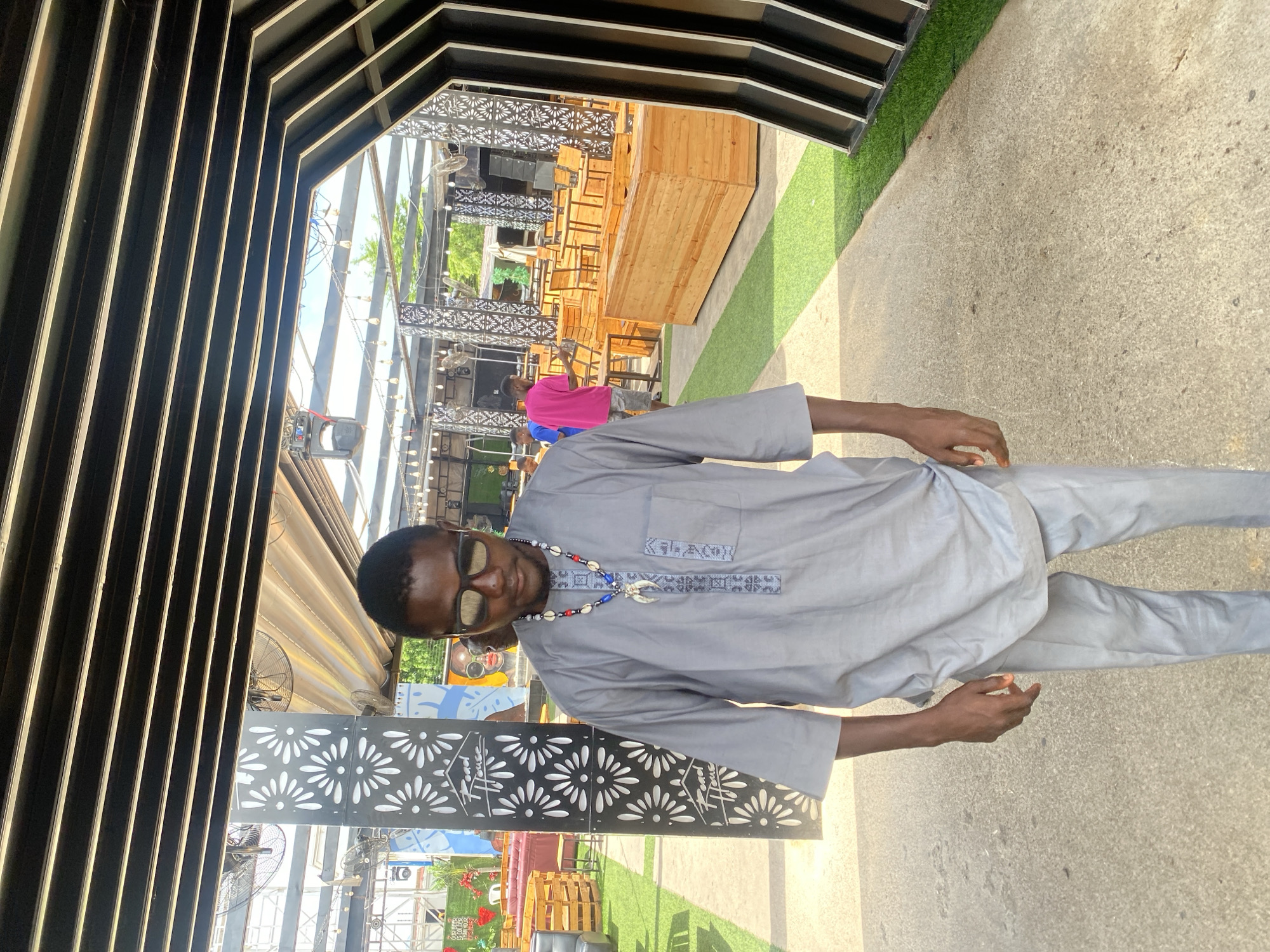 The image depicts an outdoor or semi-outdoor leisure establishment, possibly a restaurant or lounge area, located in the city of Unknown, Unknown. In the foreground, slightly left of center, stands a male with a dark complexion, wearing a grey long-sleeved tunic with matching trousers. He wears sunglasses, obscuring his eyes, and looks downwards. A prominent multi-strand beaded necklace adorns his neck, featuring red, white, and blue beads, cowrie shells, and a large white, claw-like or horn-shaped pendant. The tunic has grey embroidery detailing on the central placket and pockets.
The background reveals a structured environment with a black slatted overhead canopy, partially open to reveal a sky and green tree foliage. Some sections of the canopy have a light-colored, possibly beige, retractable fabric underneath. Industrial fans and string lights with exposed bulbs hang from the overhead structure. Decorative black metal columns with white floral or starburst cutouts are visible, one explicitly displaying "Road House" text within a logo.
To the left, there are arrangements of outdoor furniture, including red and grey upholstered lounge seating and wooden pallet-style benches. A green wall with partially visible white text, including "OUR BEER IS COLDER," is in the far background. To the right, numerous light-colored wooden tables and chairs are visible, some stacked, suggesting the area is being set up or recently used. A large wooden planter-style bench or box runs along a paved section.
Two other males are visible in the mid-ground. One wears a bright pink t-shirt and patterned shorts, standing near the wooden furniture. Another is partially visible to his left, wearing a blue shirt. The ground alternates between light grey concrete pathways and sections of bright green artificial turf. The overall scene is brightly lit by natural sunlight.