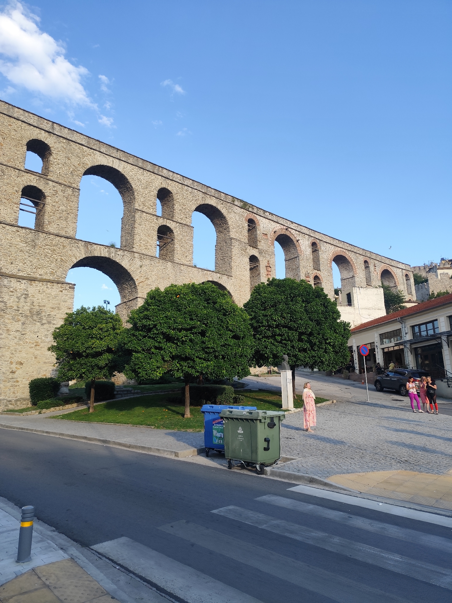 The image depicts a sunny day in Kavala, Greece, featuring a grand, ancient aqueduct as the dominant backdrop. The aqueduct, constructed from weathered stone and characterized by numerous arches, stretches across the upper portion of the frame. Below the aqueduct, a street scene unfolds. Several green trees with full foliage are planted on a grassy area, providing a stark contrast to the stone structure.
On the right side of the street, a building with a white facade and a red-tiled roof is visible, with businesses at ground level. Several people are gathered on the paved sidewalk in front of this building. Among them, three women are posing for a photo, two in casual attire and one in athletic wear. Another woman, wearing a long, light-colored patterned dress, stands further back, looking towards the aqueduct. Two large trash bins, one blue and one green, are positioned near the sidewalk. A dark-colored car is parked on the street.
The sky is a clear, bright blue with a few wispy white clouds, suggesting it is late morning or early afternoon. The sunlight casts distinct shadows, indicating a pleasant weather day. The presence of the aqueduct, the architecture of the building, and the paved cobblestone street provide strong cues about the location's historical and Mediterranean character. The overall atmosphere appears relaxed and casual, with the people engaged in everyday activities or leisure.