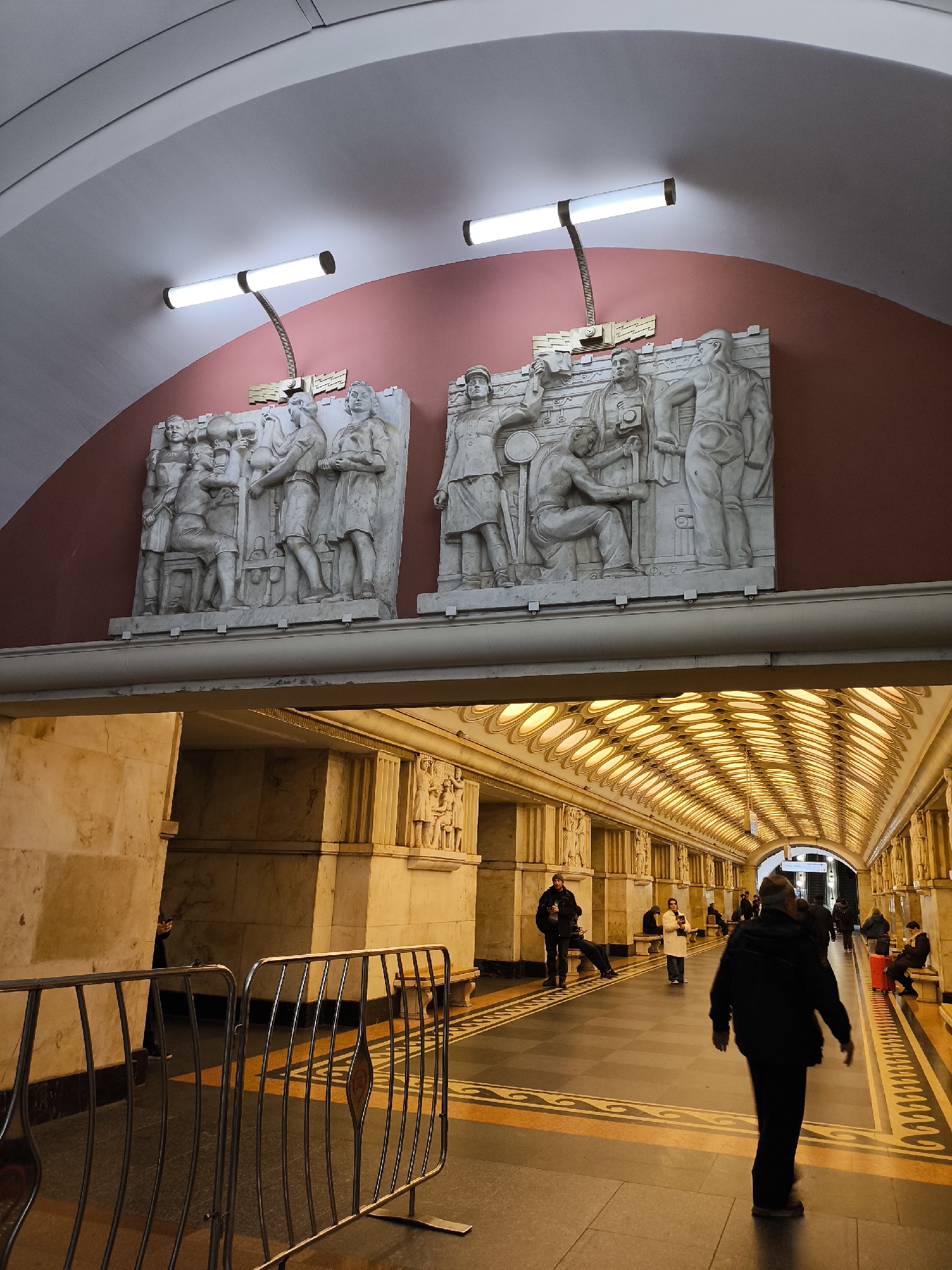 The image depicts a subway station in an unknown location. The upper portion of the image shows two bas-reliefs depicting scenes of people at work, set against a red background. Each relief is illuminated by a light fixture mounted above. Below, a wide corridor stretches into the distance. The corridor is lined with marble or stone pillars adorned with more relief sculptures. The ceiling is a grid of oval light fixtures that extend down the hallway. People are walking through the station, with one individual in a dark jacket prominently in the foreground. A metal railing is visible on the left side of the image. The floor is patterned with dark grey tiles with a light yellow border.
