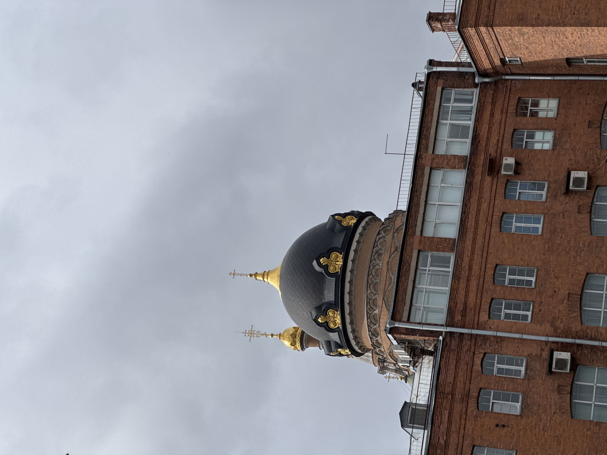 The image shows a low-angle view of a brick building and a church dome in Saint Petersburg, Russia. The brick building, likely an administrative or residential building, is on the right side of the frame and features multiple windows and air conditioning units. To the left, a dark dome with golden ornamentation and crosses marks the presence of a church. The sky is overcast and gray.