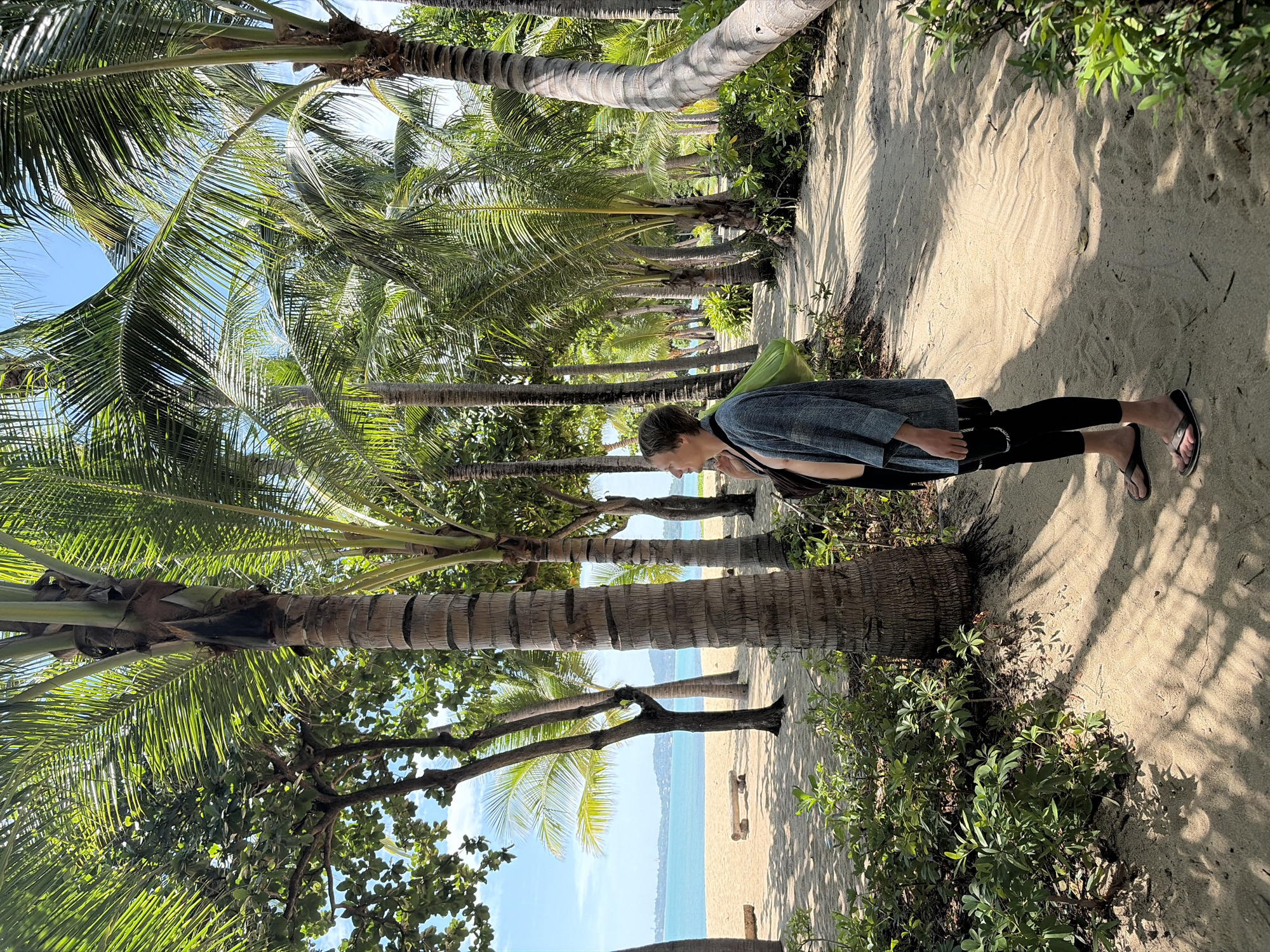 A person stands on a sandy path lined with palm trees, looking out towards the ocean. The person is wearing a light-colored open robe, black leggings, and flip-flops, with a green bag slung over their shoulder. The path is made of sand and has shadows cast by the palm trees, which are abundant and tall. In the background, there is a beach with a wooden bench, the turquoise ocean, and a distant coastline with buildings. The sky is clear and blue. The location is a beach in Ko Samui District, Thailand.