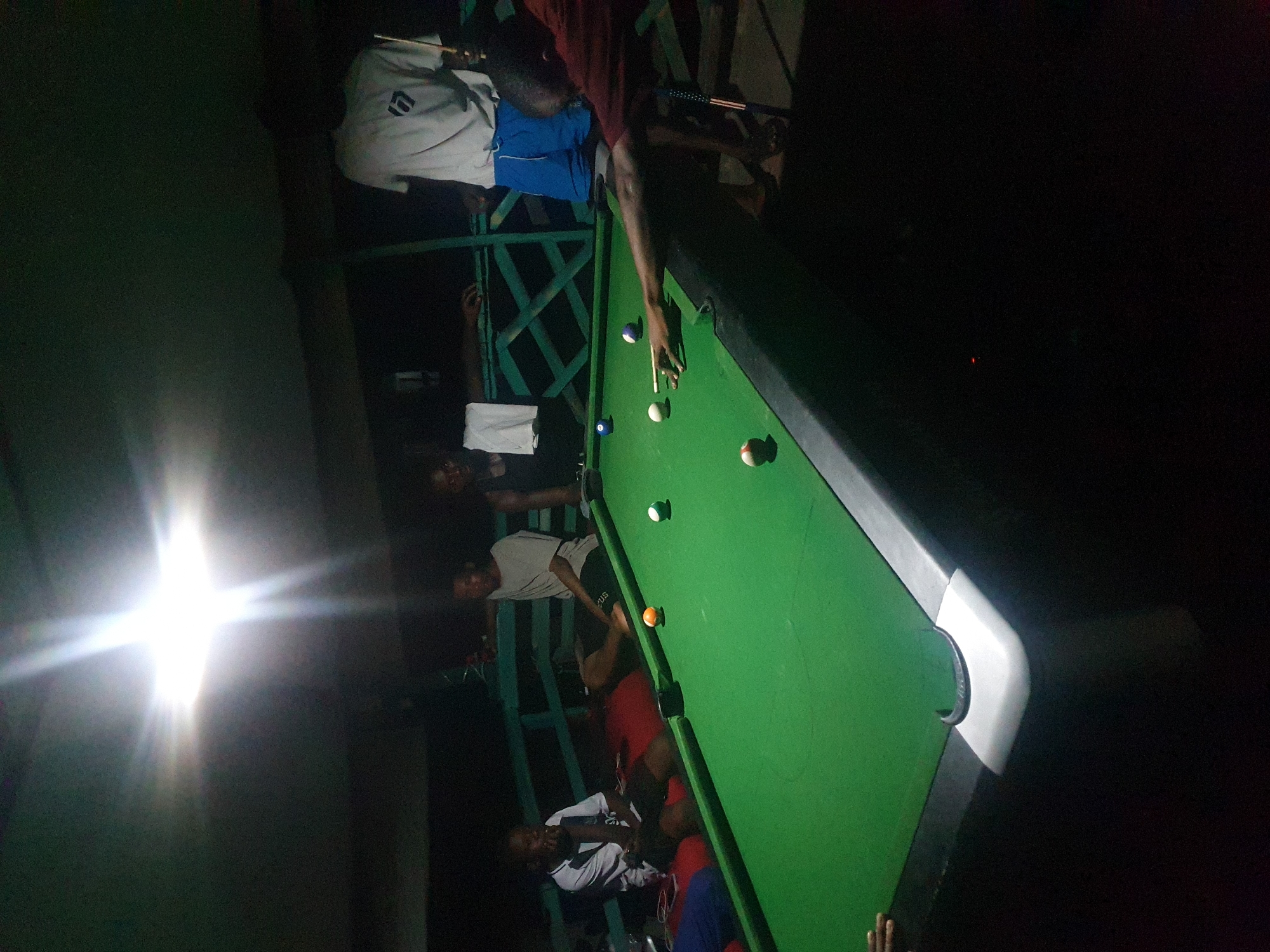 A group of young men are gathered around a pool table in an outdoor, dimly lit area. The pool table is green with various colored balls scattered across it. One man in a red shirt is actively playing, leaning over the table with a cue stick. Another man in a white t-shirt and blue shorts stands nearby, holding a cue. Several other individuals are seated or standing around the table, observing the game. The environment appears to be a casual gathering spot, possibly a bar or backyard with a covered area. The lighting is artificial, with a bright light source causing lens flare in the upper portion of the image.