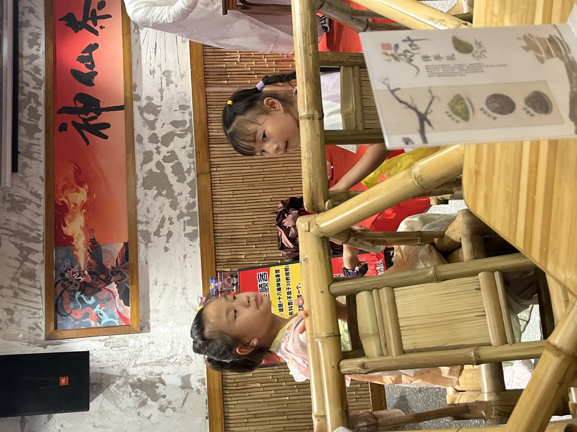 This image, taken in Yuzhong District, China, captures a candid moment of two young girls interacting in what appears to be a restaurant or tea house with a strong traditional Chinese aesthetic. The setting is characterized by bamboo furniture and decorations, including a woven bamboo wall. A prominent feature on the left is a framed artwork with Chinese calligraphy and a fiery, dynamic illustration. To the right, a menu or informational flyer is visible, displaying images of tea leaves and traditional Chinese script.
One girl, with dark hair braided and adorned with purple and yellow ties, is peering over the bamboo structure, her gaze directed downwards and slightly to the left. Her expression is one of curiosity and engagement. The other girl, also with dark hair styled in a bun, is seated and looking upwards with a thoughtful or contemplative expression. She is wearing a light-colored, possibly pink, garment with intricate detailing.
The lighting suggests an indoor setting, possibly during the day, but without strong sunlight. The overall atmosphere is warm and intimate, hinting at a family outing or a casual gathering. The visible text on the flyer includes "顾客" (customer), "神仙茶" (immortal tea), and "伴手礼" (souvenir gift), along with what seems to be a promotional tagline mentioning "十八梯神仙茶馆" (Shibati Immortal Tea House). The imagery and text collectively point to a place that celebrates traditional Chinese tea culture.