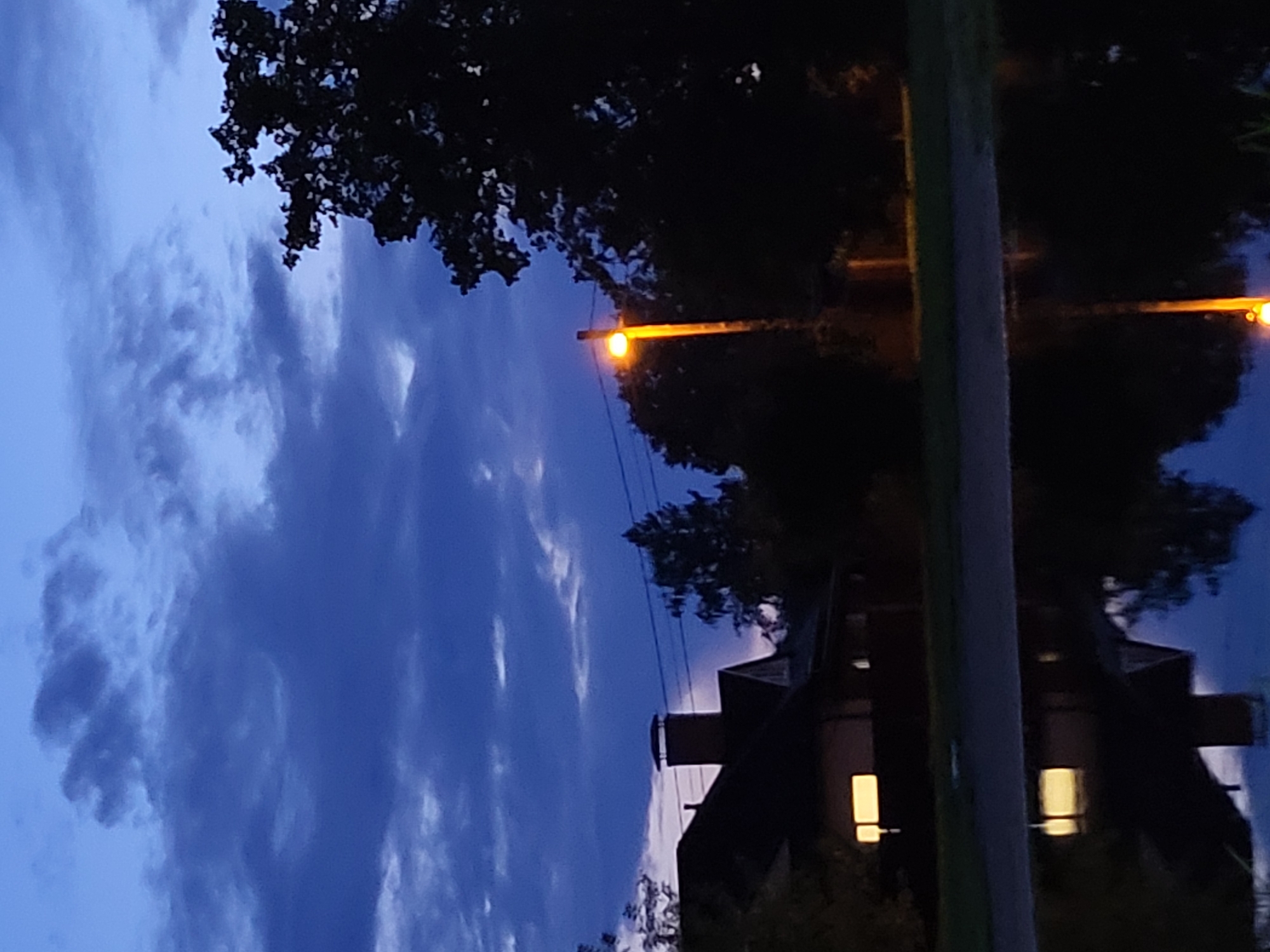 The image shows a twilight scene in Jelgava, Latvia, with a dark blue sky filled with textured clouds. In the foreground, a large puddle or body of water reflects the sky and the surrounding environment. To the left, a silhouetted house with lit windows and a prominent chimney is visible. A street lamp with a bright orange glow stands to the right, partially obscured by a large, dark tree. The overall atmosphere is serene and quiet, characteristic of the evening hours in a residential area.