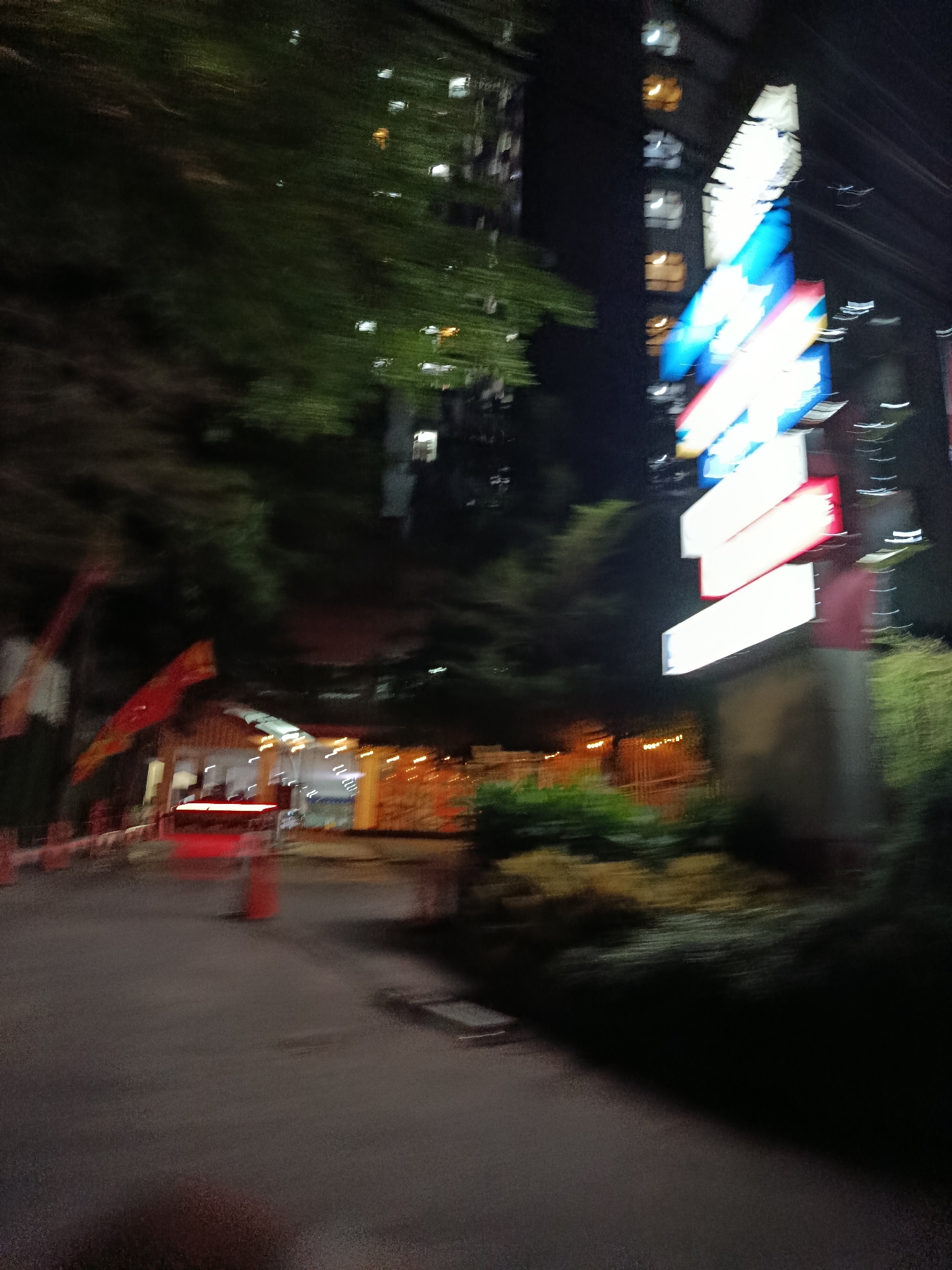 The image captures a blurred nighttime street scene in Depok, Indonesia. In the foreground, a dark asphalt road leads towards a small structure with a red, illuminated canopy, possibly a food stall or shop. Several orange traffic cones are visible to the left of the road. Further back, a tall building with illuminated windows looms in the background. To the right, a stack of brightly colored, illuminated signs, predominantly white and blue, stands prominently against the dark sky. Green foliage is present on the right side of the frame, partially obscuring the building. The overall impression is one of a bustling urban environment at night, characterized by artificial lighting and motion blur, suggesting movement of the camera or a fast shutter speed.