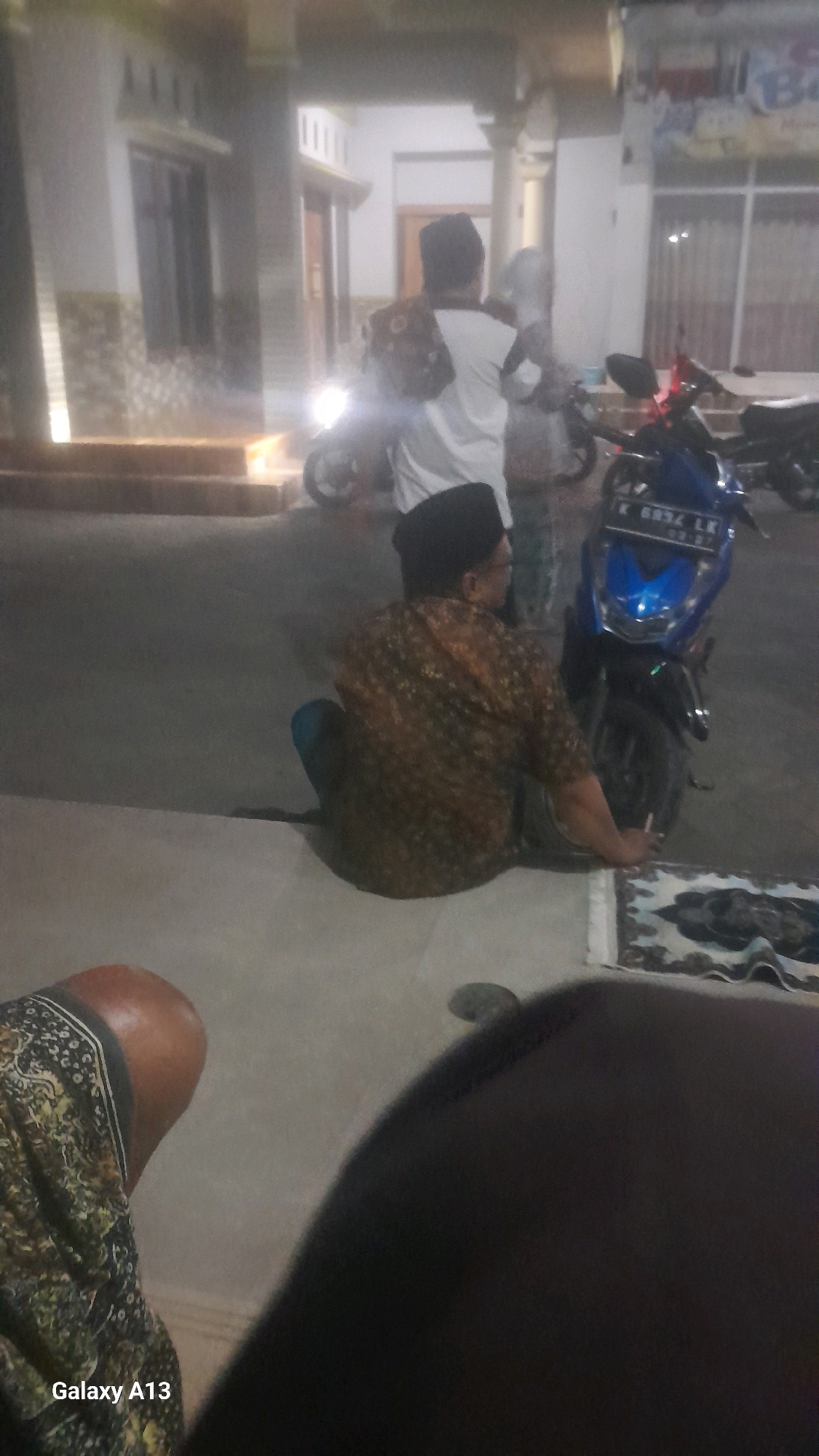 In the city of Ngelo, Indonesia, at night, two men are visible. One man, wearing a black peci and a patterned brown shirt, sits on the ground. Another man, wearing a white shirt and peci, stands in the background, carrying a bag. A blue motorcycle is parked nearby. The scene appears to be in front of a building, possibly a house, with a well-lit entrance. A prayer mat is placed on the ground in front of the sitting man. The overall lighting suggests it is evening or night.