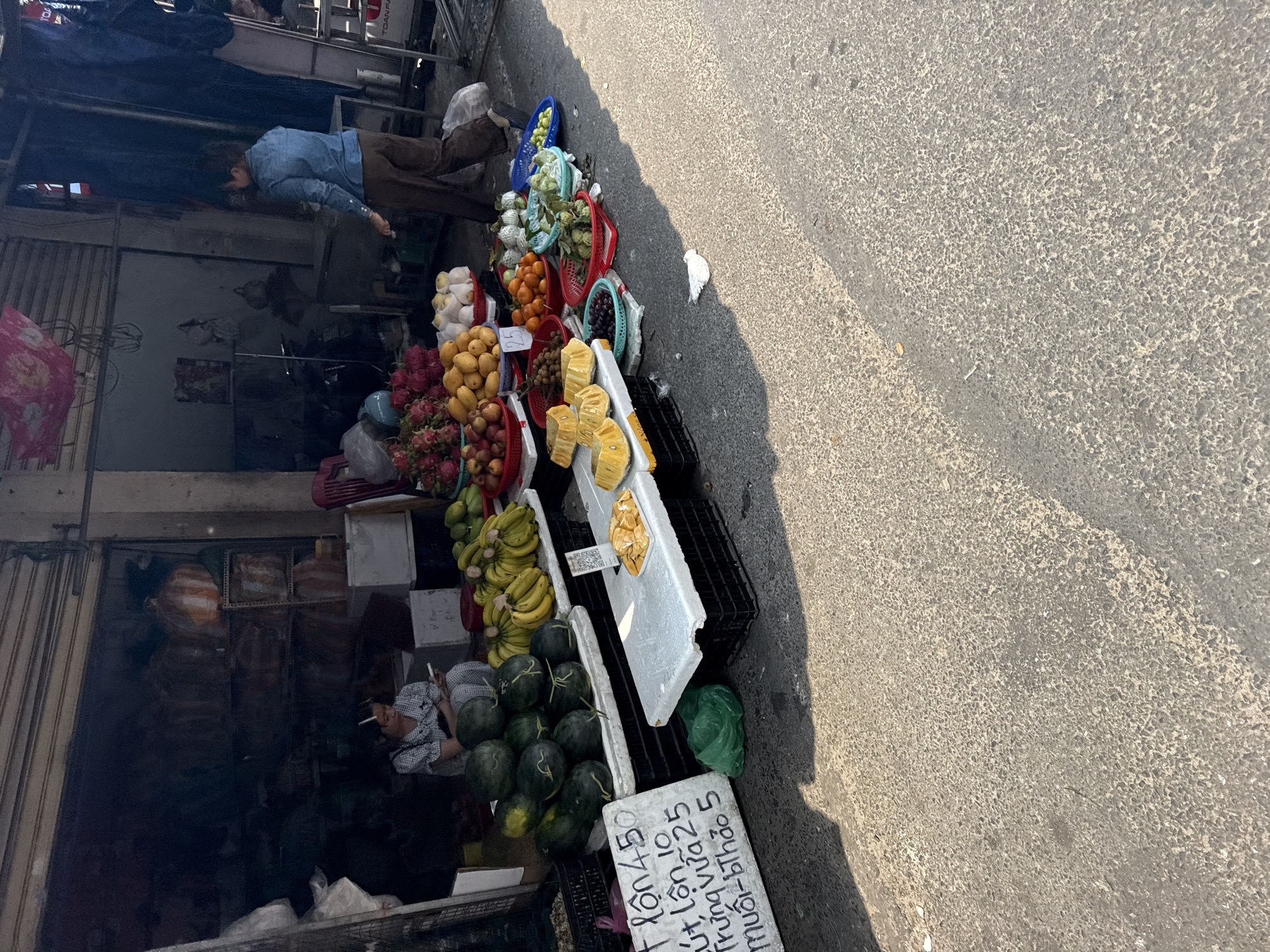 A street scene in Đà Nẵng, Vietnam. Fruit is displayed for sale on the sidewalk next to a building. Fruits such as watermelons, bananas, dragon fruit, mangoes, tangerines, guavas and pineapples are visible. A sign on a white board lists prices. A man is standing near the fruit stand, looking toward the fruit. Another man is lying down inside the shop and smoking. The street and building cast shadows.