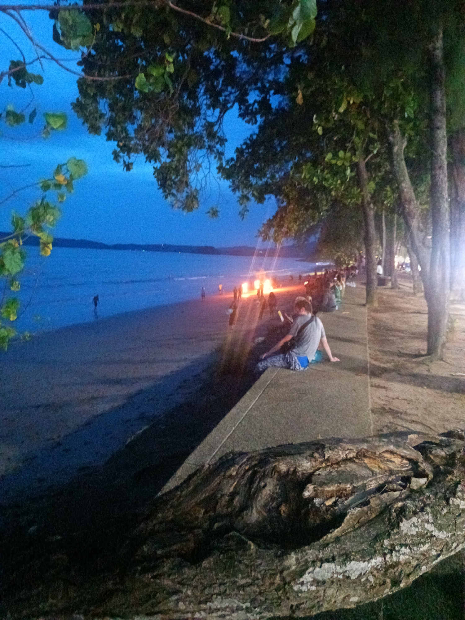 The image depicts a nighttime scene at Ban Ao Nang, Thailand. The scene includes a beach with a body of water in the background, and people, likely tourists, gathered around a bonfire on the beach. The image is taken from a higher vantage point, with a man sitting on a low wall. Another row of people are sitting further down the walkway. Trees line the walkway adjacent to the beach. A large piece of driftwood is visible in the foreground. The sky is a dark blue, suggesting twilight. Streetlights can be seen behind the row of trees.