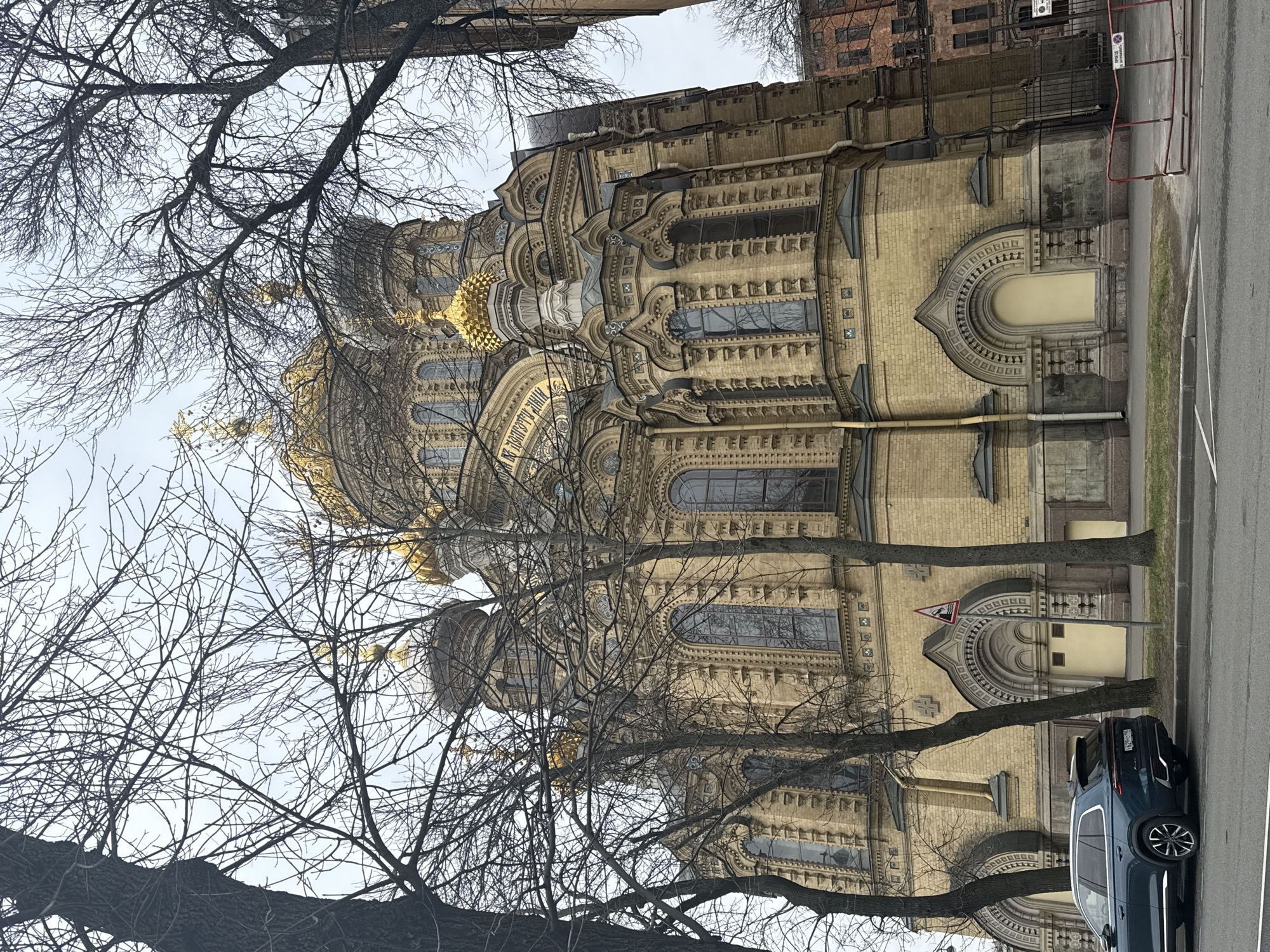 The image displays a grand, ornate church-like building, likely a cathedral, characterized by its intricate architecture and golden domes, set against an overcast sky. The building is constructed of light yellow brick or stone, featuring numerous arched windows, many with stained glass patterns, and richly detailed facades. Multiple golden, onion-shaped domes and cupolas gleam at the top, reflecting the ambient light. Decorative elements include ornate cornices, pilasters, and carved reliefs, contributing to its majestic appearance.
In the foreground, bare trees with dense, leafless branches partially obscure the building, particularly its upper sections and domes. These trees suggest a late autumn, winter, or early spring season.
A paved street runs along the right side of the building, with a sidewalk and curb clearly visible. A dark blue SUV is parked on the street. A red and white triangular warning sign depicting a road bump is prominently placed near the street. Another small, illegible sign is affixed to a fence in the background.
Visible Cyrillic script can be partially deciphered on the building's facade, specifically on a large arched relief. Phrases such as "ЧЬЕ ВЫСОКО" and "ВЛАСТЬЮ И ЦАРЯ" are discernible. The sky is uniformly overcast, providing diffused daylight, and there are no people or specific activities taking place in the scene. The overall impression is one of architectural splendor framed by nature's starkness.
