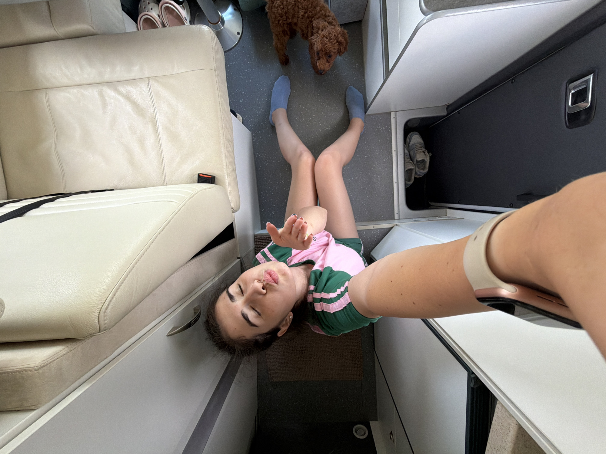 The image shows a young woman and a dog inside a vehicle, possibly a camper van or RV. The woman is taking a selfie, puckering her lips as if to blow a kiss. She is wearing a pink and green t-shirt and blue slipper socks. The dog, a small, curly-haired breed, is lying on the floor next to her. The interior of the vehicle appears to be upholstered in light-colored leather and has a practical, utilitarian design. There are storage compartments visible, one of which contains a pair of shoes. The flooring is a dark gray speckled material. The image is shot from a high angle, looking down on the woman and dog. The scene is well-lit, suggesting it is daytime. The overall impression is one of a casual, indoor setting within a vehicle. There is no visible indication of the city of Nea Moudania, Greece, in this image.