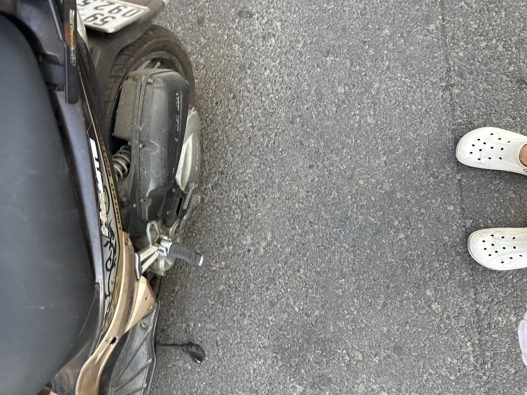 The image shows a portion of a black and gold scooter parked on a grey asphalt surface. To the right of the scooter are a pair of white Crocs worn by a person whose lower legs are partially visible. The location is likely Bac Nha Trang Ward, Vietnam, based on provided information. The scene appears to be a street or parking area.