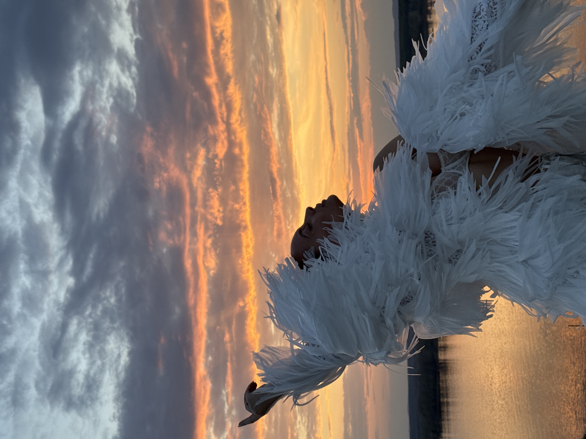 A woman in a feathered white garment is oriented horizontally against a sunset backdrop with water in the lower right corner. The sky presents a mix of blue-gray clouds and orange-yellow hues. The location is unknown, and there is no evidence to indicate this is in the city of Ольховка, Russia.