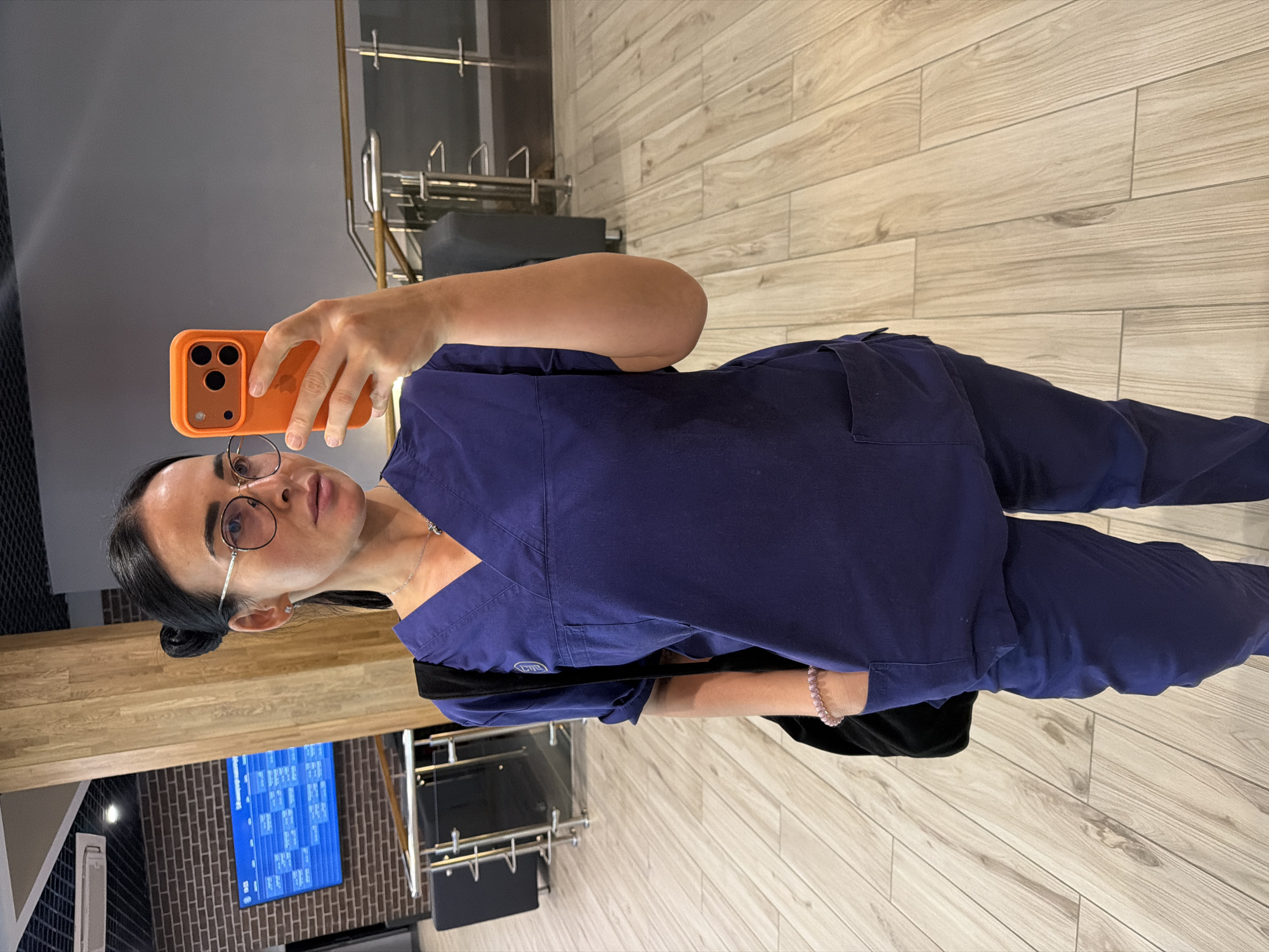 A woman with dark hair pulled back in a bun and wearing clear-framed glasses stands upright, taking a mirror selfie. She is dressed in dark blue medical scrubs, consisting of a v-neck top with pockets and matching pants. A silver necklace with a small pendant is visible around her neck, and a light pink beaded bracelet is on her left wrist. She holds an orange-cased iPhone with three camera lenses in her right hand, positioned in front of her face, with the Apple logo partially visible on the phone case. Her expression is neutral, and she is looking directly into the phone's camera, reflecting off a mirror.
The setting appears to be an indoor commercial or professional space, possibly a clinic or waiting area, in Saint Petersburg, Russia. The background features light-colored wooden floorboards laid in a horizontal pattern. Behind her, a dark brick accent wall is visible, with a blue digital screen displaying what looks like a schedule or information grid in white text. To the right, there are metallic railings and what seems to be dark furniture or seating. Another reflective surface is to her left. The overall environment is modern and well-lit by unseen indoor lighting. No specific time of day is discernible. A small, indistinct logo is visible on the right chest pocket of her scrub top.