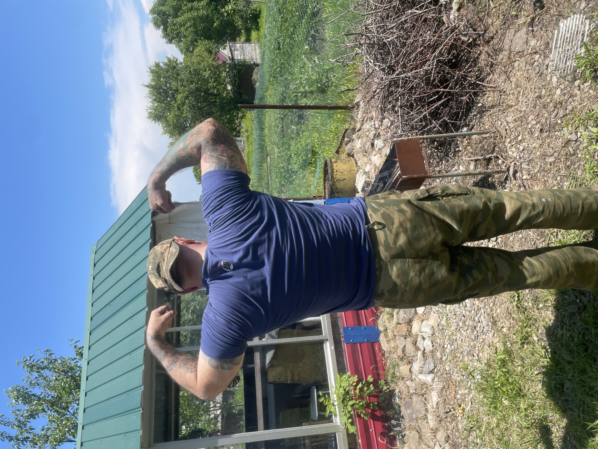 An outdoor, eye-level, medium shot captures a Caucasian man, viewed from behind, flexing his biceps under a bright blue sky with scattered white clouds. He wears a camouflage baseball cap, a dark blue t-shirt with a small black logo on the back, and camouflage cargo pants. His arms are covered in colorful tattoos.
The man stands on a gravelly area in front of a red-sided structure with a green, corrugated metal roof. To his left is a greenhouse-like structure with glass panels. Behind him, a wire fence encloses a green field of crops, with a rustic wooden building visible in the distance. A rusty barbecue grill with burning embers sits to the right of the man, accompanied by a large pile of dry branches and twigs. The overall setting suggests a rural or suburban environment, possibly a backyard or garden area. The scene is well-lit by natural sunlight, indicating a daytime shot.