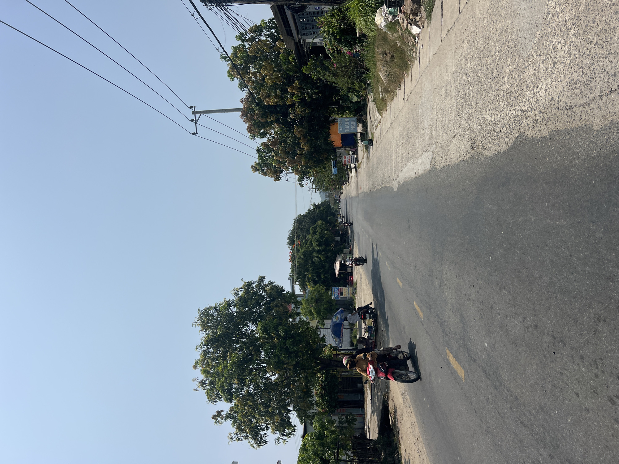 The image depicts a street scene in Bắc Nha Trang, Vietnam. A paved road stretches into the distance, lined with trees on both sides. Overhead power lines are visible against a clear sky. Several motorbikes are present on the road and parked along the roadside. Some buildings and signs are partially visible amongst the trees. The lighting suggests it is daytime. The road surface appears to have yellow dividing lines.