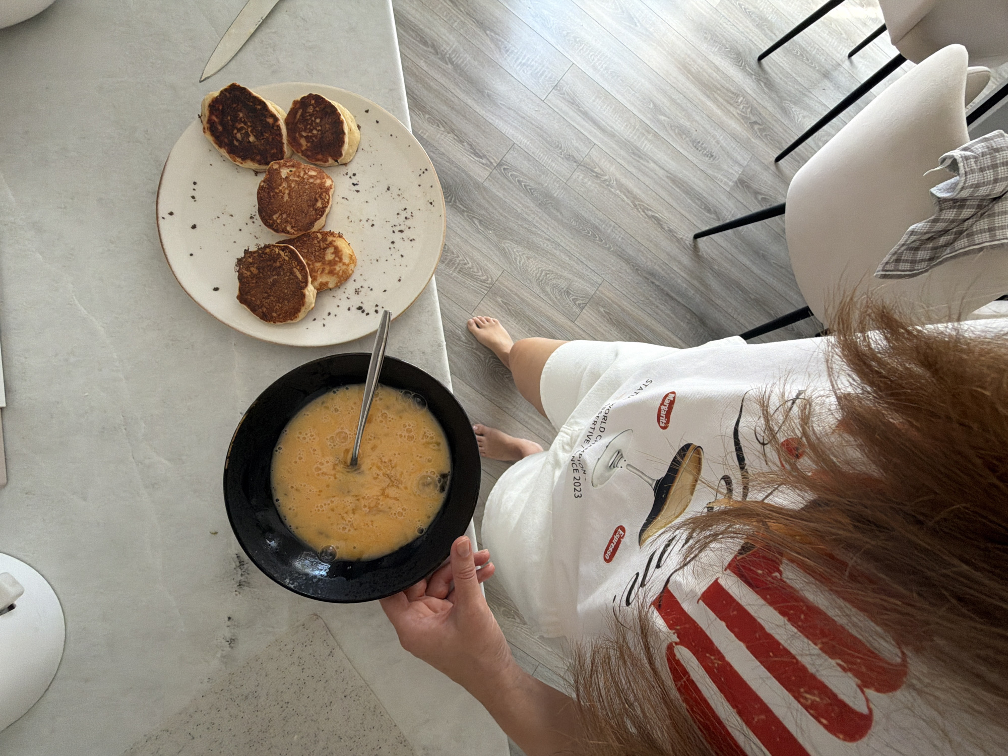 A top-down view of a person preparing pancakes in what appears to be a kitchen in Malinovka, Russia. The person is wearing a white t-shirt with a red and white striped design and images of drinks, including a martini glass and the word "Margarita." They are holding a black bowl filled with whisked eggs, with a metal whisk submerged in the mixture. On a white plate, six small, golden-brown pancakes are arranged, with some dark specks, possibly pepper or spices, scattered around them. A silver knife is positioned to the right of the plate. The background shows a light-colored countertop and a wooden floor with a greyish tone. Two chairs with light-colored upholstery and black legs are visible in the lower right corner. The overall scene suggests a casual breakfast preparation.