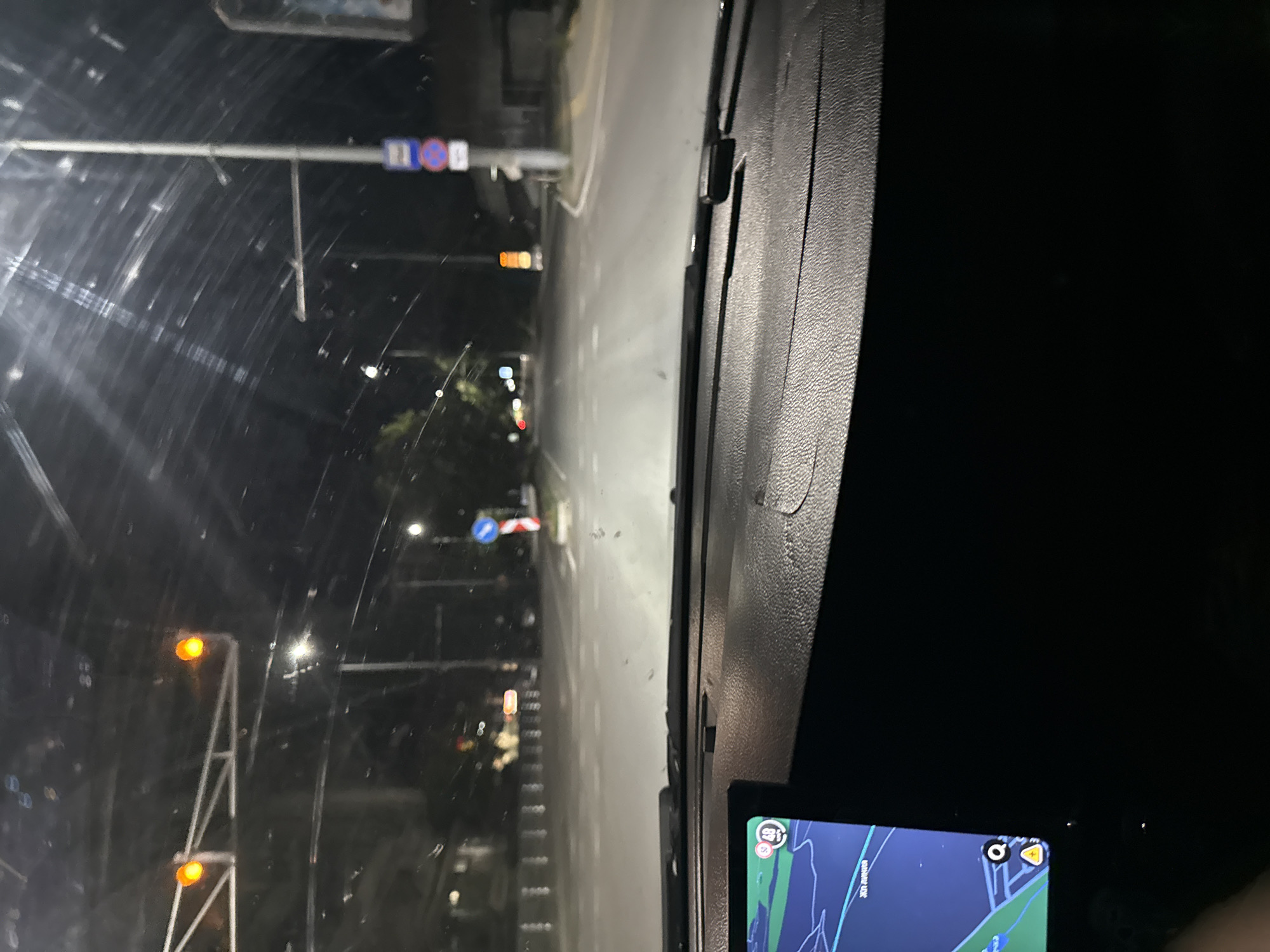 The image depicts a night-time scene viewed from inside a vehicle, likely from the driver's perspective. The windshield shows a wet or dirty surface, with streaks of light and reflections. Outside, a road is visible, illuminated by streetlights and the headlights of the vehicle. Traffic signals and signs are present, including a blue and white sign and a red and white sign. On the left side of the frame, there are what appear to be overhead structures with lights, possibly streetlights or part of a building. Further down the road, more lights are visible, suggesting an urban or suburban environment.
The right side of the image shows the interior of the vehicle, with a textured dashboard. A GPS device or phone screen is visible in the lower right corner, displaying a map interface with routes and possibly navigation information. Some text is visible on the GPS screen, but it is not clearly legible.
The overall impression is that of driving at night, possibly in rainy conditions due to the streaks on the windshield. The presence of road infrastructure and the GPS device indicates travel. The time of day is clearly night, and the lighting suggests an outdoor, potentially urban setting in Stara Zagora, Bulgaria. There are no people clearly visible, and the focus is on the road ahead and the vehicle's interior.