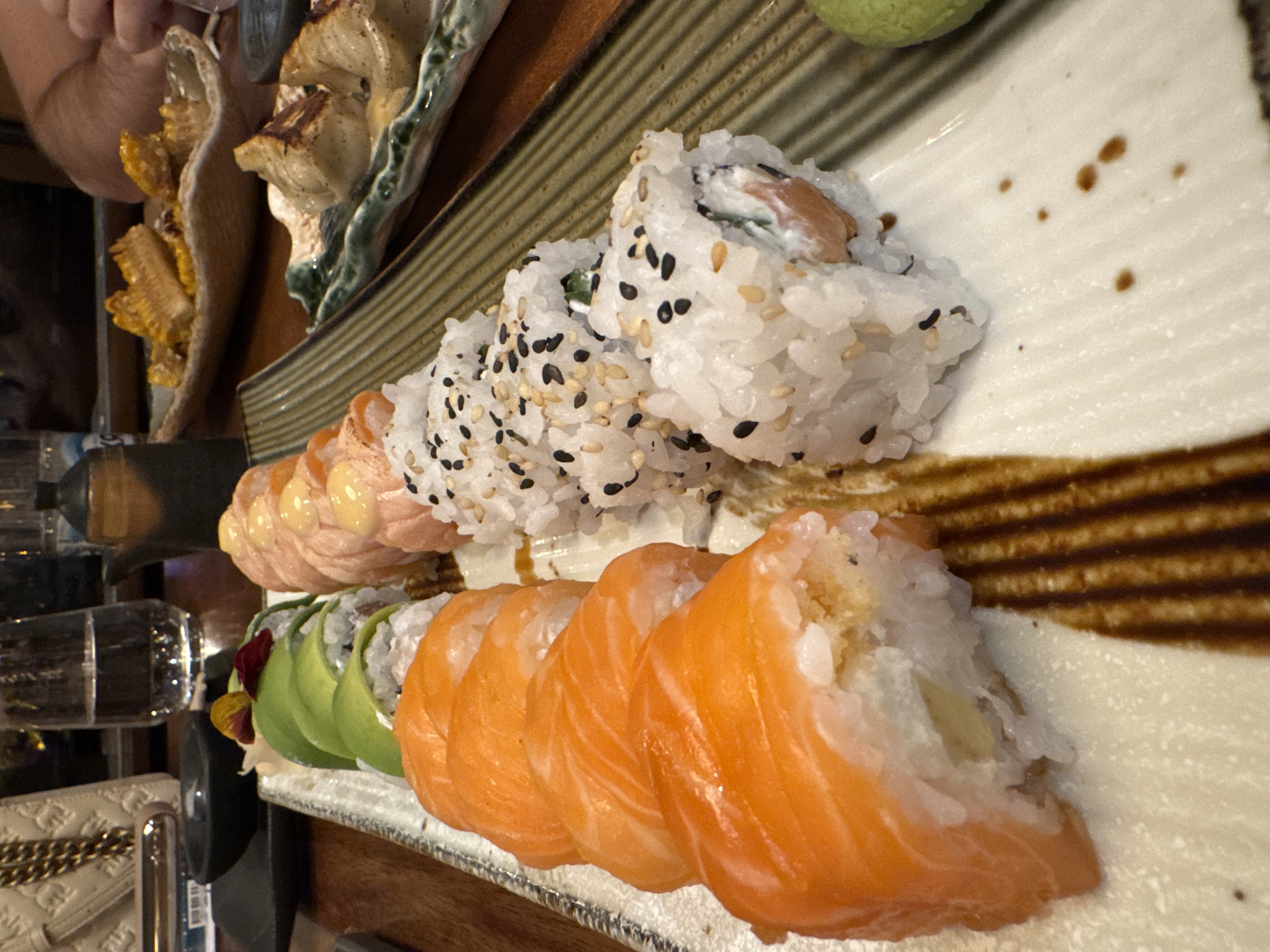 A close-up, medium-angle shot shows a platter of sushi rolls. The sushi is arranged in a line, consisting of several varieties including a salmon-topped roll, a sesame-seed coated roll, and an avocado roll. There are other dishes out of focus in the background.
The scene appears to be indoors, possibly at a restaurant. The location is implied to be Sofia, Bulgaria. No people are clearly visible in the primary focus of the image, though part of a person's arm is visible in the upper left corner. The overall environment seems to be a restaurant setting.