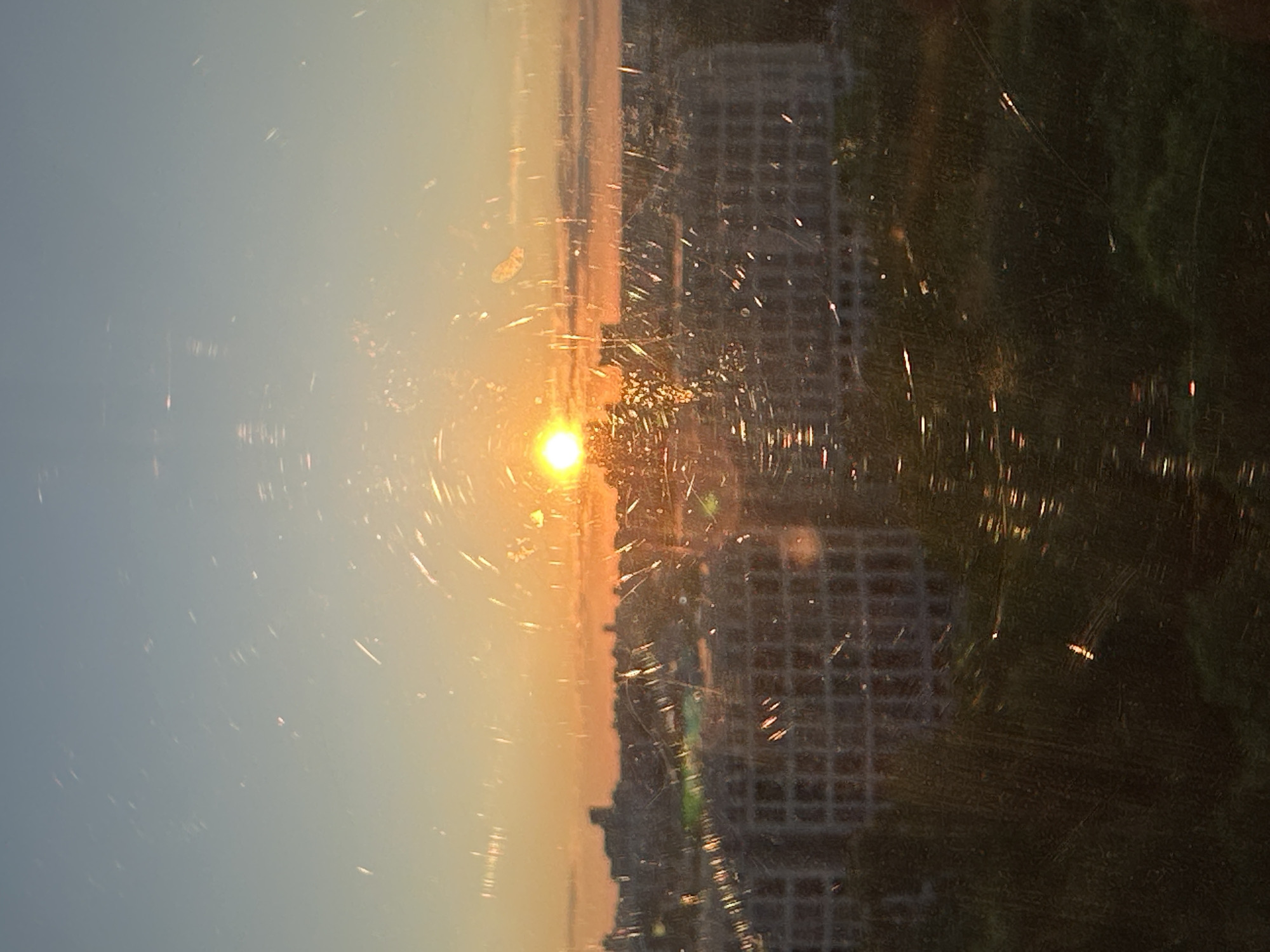 The image captures a vibrant sunrise or sunset viewed through a heavily scratched and dusty window. The scene is oriented vertically in the original, but mentally rotating it 90 degrees clockwise presents a standard landscape.
The sky dominates the upper and left portions of the frame (after rotation), transitioning from a pale, clear blue to a warm, glowing orange and yellow closer to the horizon. A brilliant, circular sun is the focal point, positioned low on the horizon, emitting intense light.
Below the sun and against the radiant sky, a dark silhouette of a cityscape is visible, featuring various buildings with discernible rectangular window patterns. These structures form a distinct horizon line.
The immediate foreground is dominated by the window pane itself, which is extensively covered in scratches, streaks, and specks of dust or moisture. These imperfections are brightly illuminated by the sun's rays, creating numerous scattered points of light and lens flare effects, especially prominent in the areas directly in front of the sun and extending across the right side of the image. The texture of the window glass partially obscures the view of the city and a darker, possibly green, area in the lower right, which could be vegetation or a darker part of the urban landscape.
The image depicts the peaceful and dramatic moment of either dawn or dusk, observed from an interior vantage point. The scratches on the glass add a raw, unfiltered layer to the viewing experience. There is no visible text in the image. While the location is provided as Minsk, Belarus, the visual content does not contain specific identifying landmarks for the city.