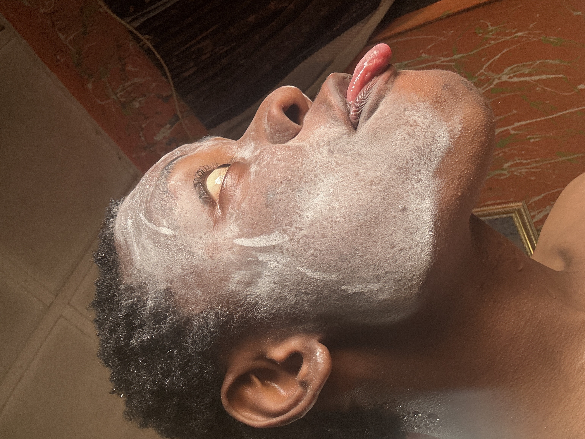 The image depicts a close-up, upward-angled view of a person's face and neck, covered with a white substance resembling a facial mask or powder. The person's mouth is open, with their tongue extended upwards. The person's skin is dark. Their hair is short, dark, and curly. The right ear is visible in the lower left of the image. The person's right eye is open.
The background consists of a wall with an orange painted finish. The upper left of the image shows an unpainted part of the wall where you can see red bricks and black materials. A gold frame is visible in the lower right corner.
No specific information regarding the location or city is discernible from the image.