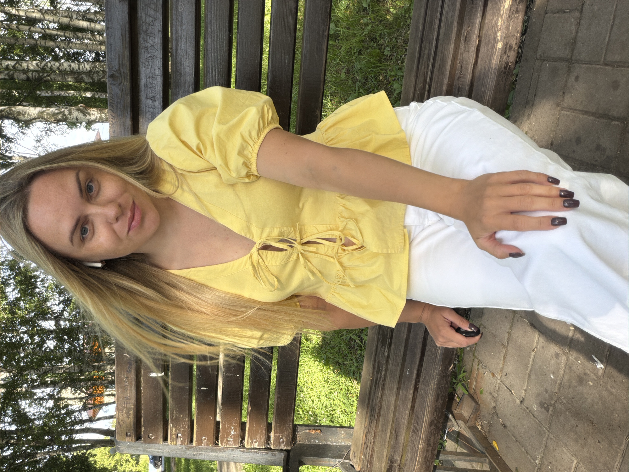 A young woman with long blonde hair is sitting on a wooden park bench. She is wearing a yellow top with puffy sleeves and white pants. The woman is looking at the camera and smiling. Her left hand is resting on her lap, and her right hand is holding a small black object, possibly a phone or a remote. She is wearing white earbuds. The background shows green grass and trees, suggesting an outdoor setting in a park. The sun appears to be shining, casting shadows on the bench and the paved walkway next to it. The image orientation is rotated 90 degrees clockwise. The scene suggests a relaxed and pleasant day outdoors in Votkinsk, Russia. There is no visible text in the image.