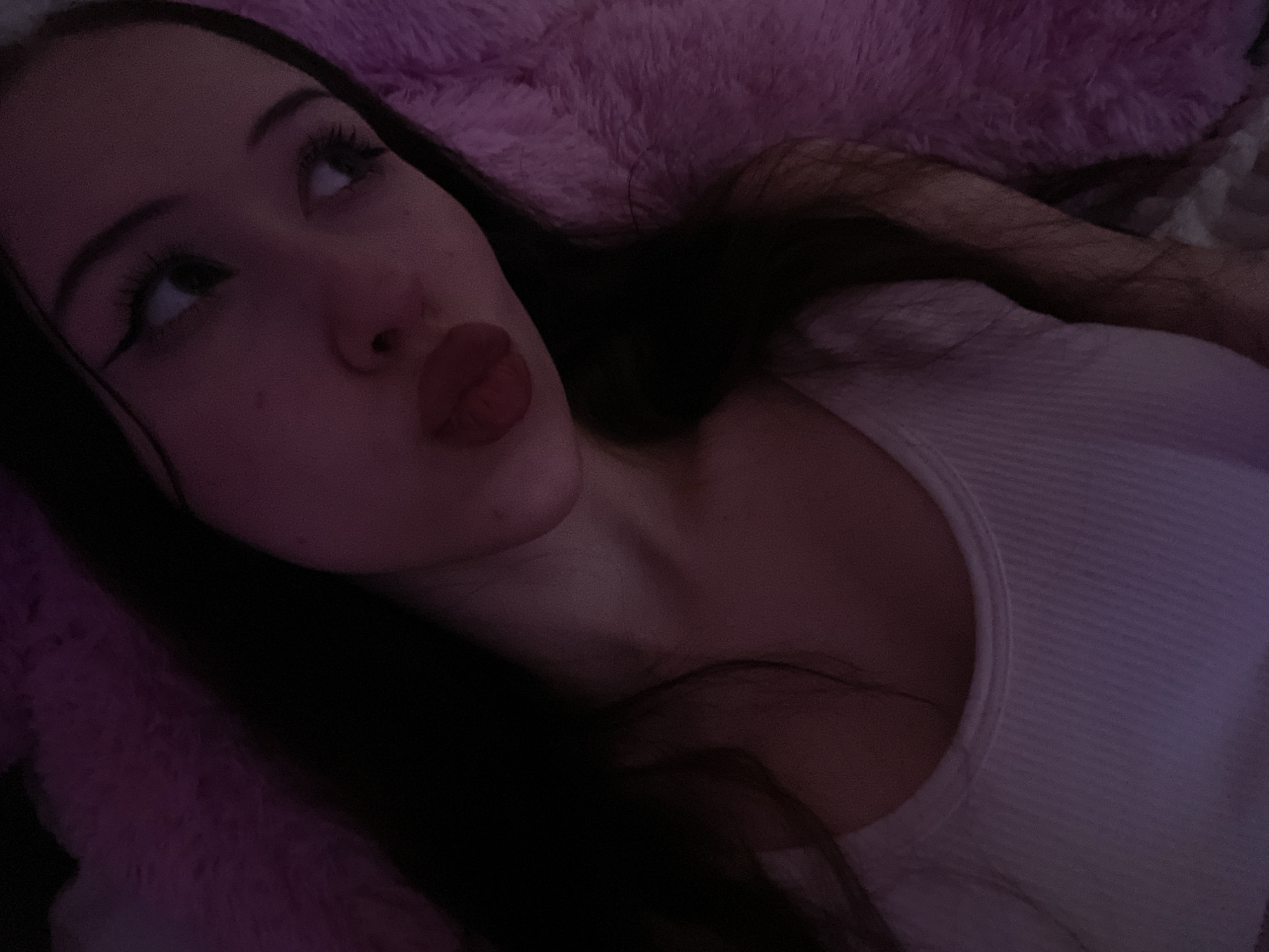 The image shows a close-up, selfie-style photograph of a young woman lying down. She has long, dark hair and is wearing a white tank top. Her makeup includes winged eyeliner. She is looking off to the right side of the frame with her lips pursed. The background is out of focus and appears to be a soft, textured, pink surface, possibly a blanket or fur. The lighting is dim and has a pinkish hue. There is no indication of the specific location within Votkinsk, Russia.