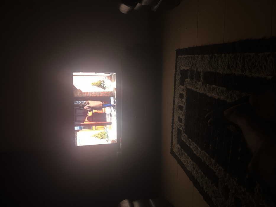 A dark room with a brightly lit television screen showing a person standing in front of a house. To the right is a dark rug on the floor with a geometric pattern. In the bottom right corner, part of a person's leg and foot is visible. Some objects are on the top of the TV and in the bottom of the image, but they are difficult to make out due to darkness.