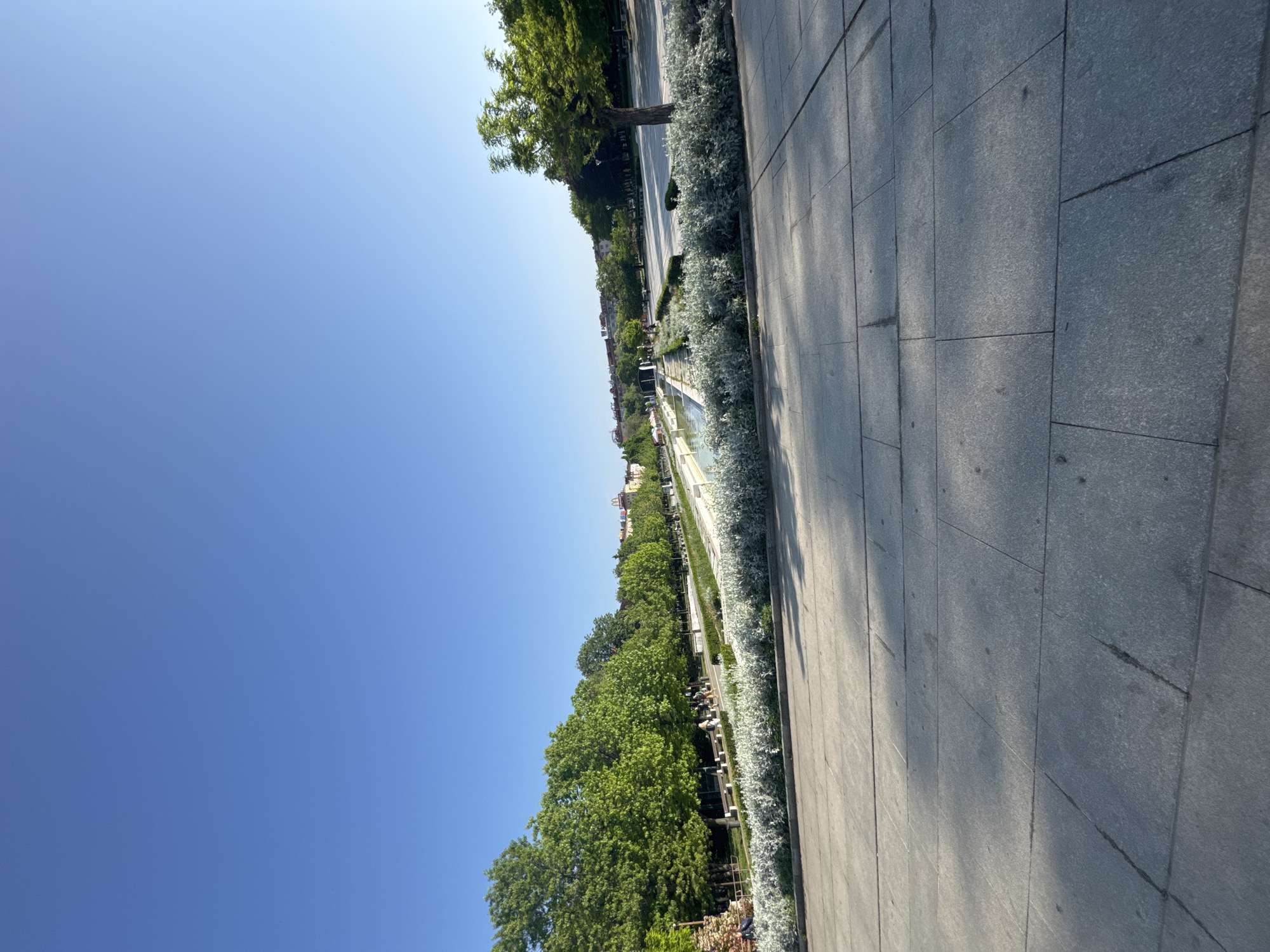 The image captures a sidewalk on a sunny day in Sofia, Bulgaria, near a landscaped canal. The sidewalk is paved with large, grey rectangular stones. Adjacent to the sidewalk is a border of silver-leafed plants, followed by trees and benches lining the canal. In the distance, buildings are visible. The sky is clear blue.