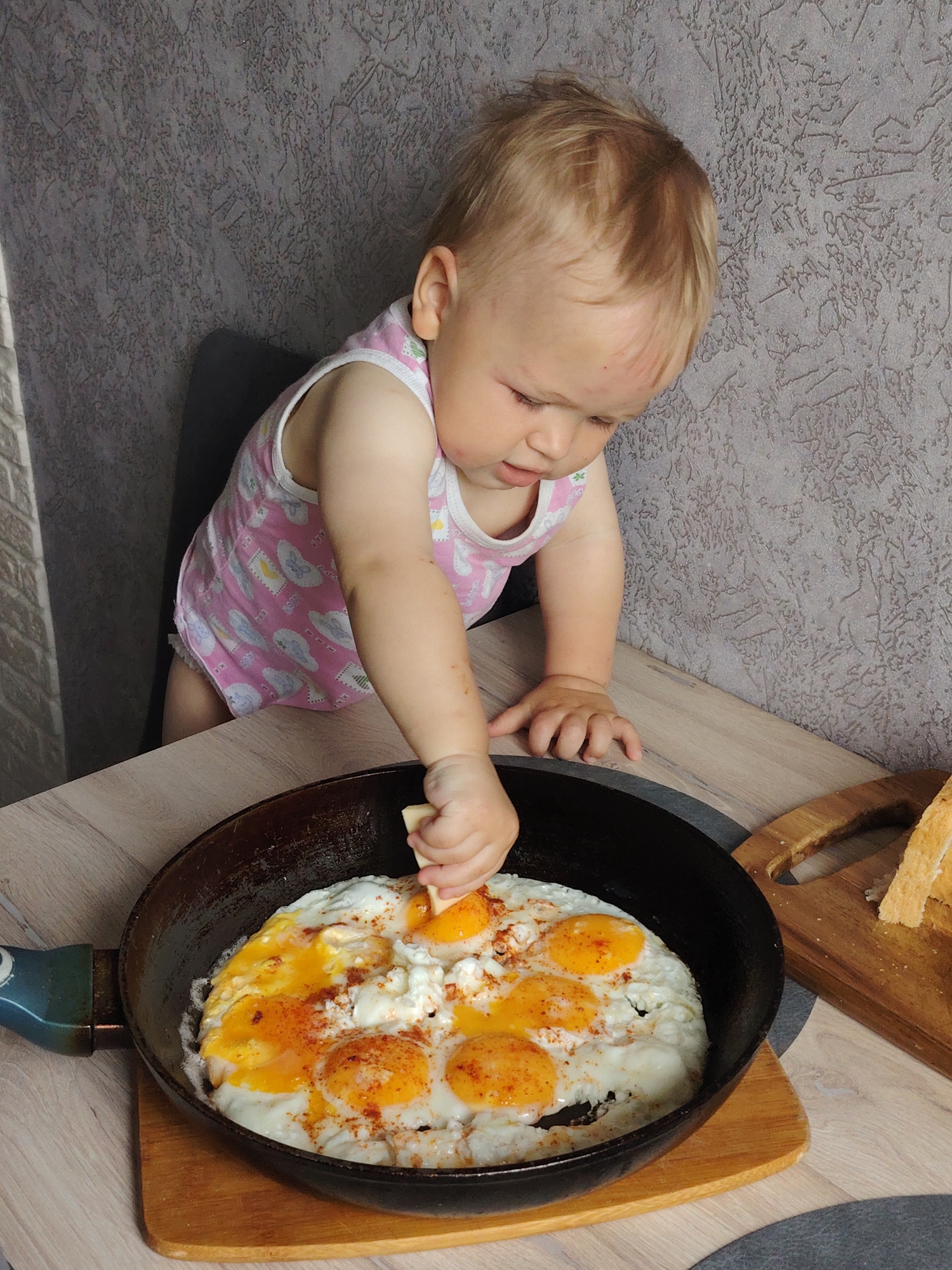 In a room with gray wallpaper and a light-colored wooden table, a toddler wearing a pink onesie with white designs stands on a chair, reaching towards a black frying pan filled with several fried eggs with bright orange yolks. The toddler's right hand holds a piece of bread or cheese, poised above the eggs. A wooden cutting board with slices of toast sits next to the pan. The photo was taken in the city of Chaikovsky, Russia.