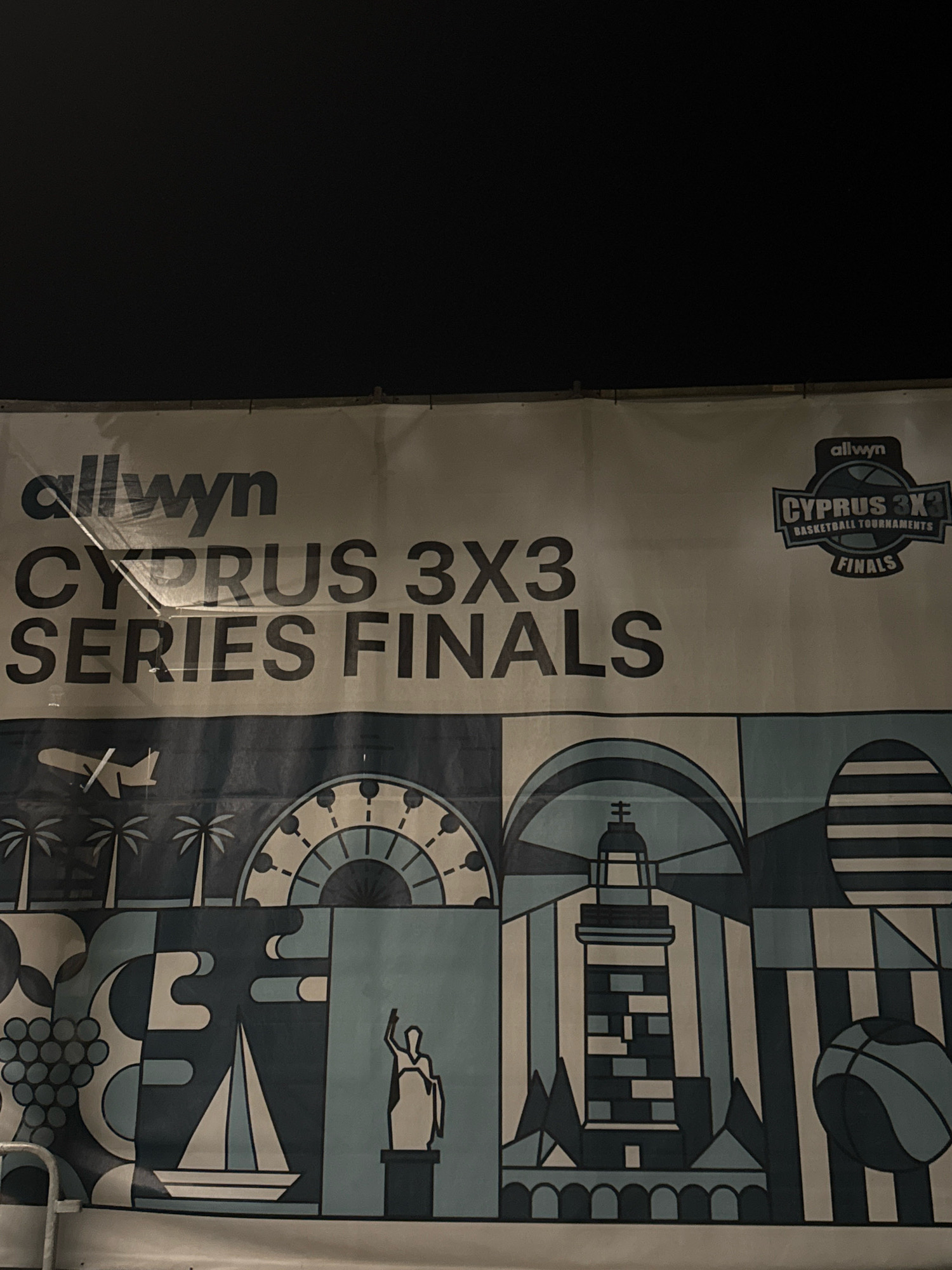 The image shows a banner for the "allwyn CYPRUS 3X3 SERIES FINALS BASKETBALL TOURNAMENTS". The banner is light-colored with large, dark lettering. The top portion of the banner has the text "allwyn" in smaller font on the left and a logo with "allwyn", "CYPRUS 3X3 BASKETBALL TOURNAMENTS", and "FINALS" on the right. The main text reads "allwyn" followed by "CYPRUS 3X3 SERIES FINALS". The bottom portion of the banner features a graphic design with stylized images of elements associated with Cyprus, including palm trees, a Ferris wheel-like structure, a sailboat, a lighthouse, and a basketball. The image was taken at night, as indicated by the dark sky. The banner appears to be displayed outdoors, possibly in a public area or at a sporting event venue. Based on the text, the event is taking place in Cyprus. The graphic elements suggest a coastal or Mediterranean setting, which is consistent with Limassol being a coastal city in Cyprus.