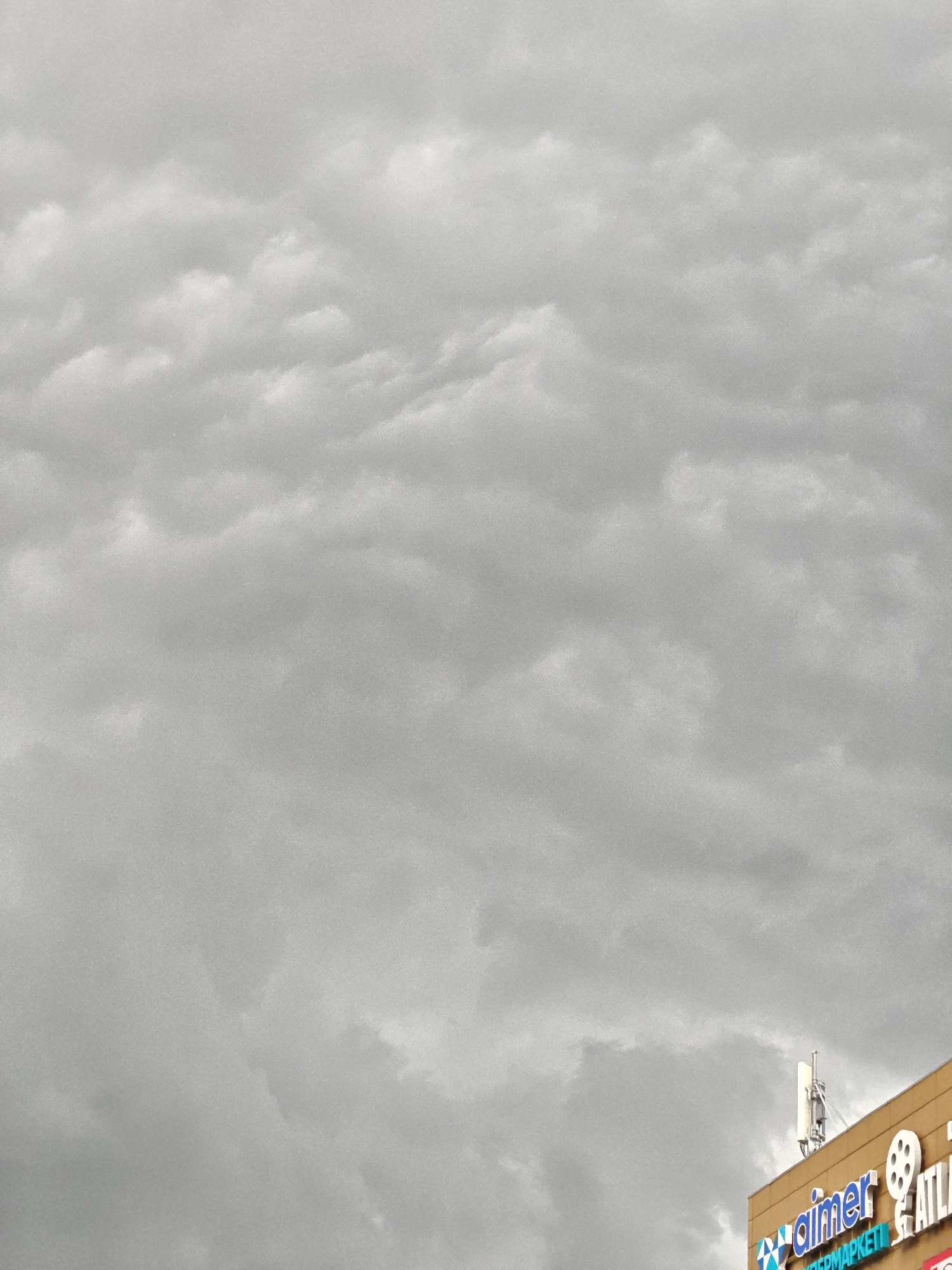 The image shows a cloudy sky over a building in Petropavl, Kazakhstan. The building has a sign that reads "aimer" in blue letters and a film reel logo. Below "aimer", the word "SUPERMARKET" is partially visible in blue letters. A white antenna is mounted on the roof of the building. The sky is overcast with gray clouds.