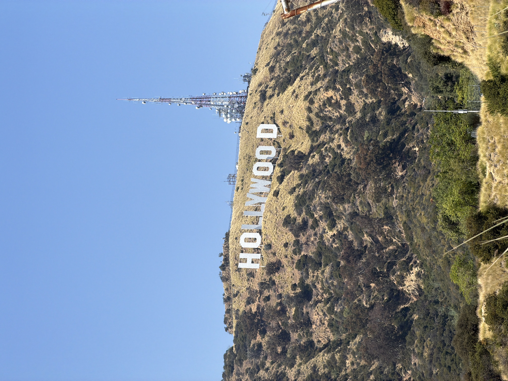 This image showcases the iconic Hollywood sign, a landmark located in the Hollywood Hills area of Los Angeles, California. The sign is displayed in large, white capital letters along the slope of a mountain. A communications tower can be seen on the ridge behind the sign. The terrain is rugged, with vegetation consisting of dry grass, brush, and trees. The sky is a clear, bright blue.
