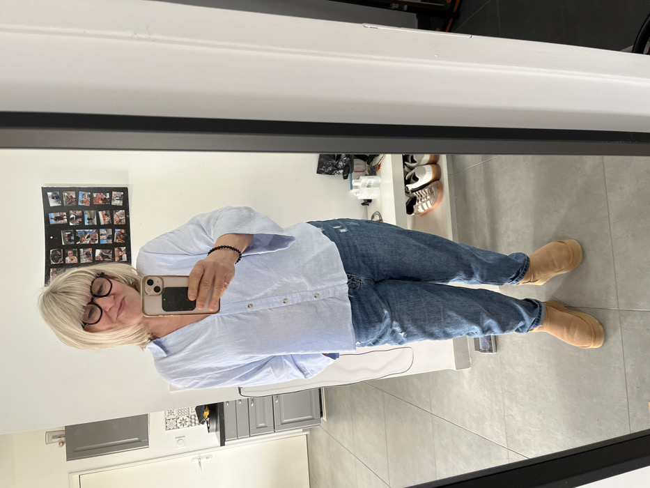 This image, situated in Chambœuf, France, depicts an adult woman captured in a full-length mirror selfie. The woman has short, layered blonde hair, wears black-framed eyeglasses, and exhibits a subtle smile while holding a light-colored smartphone with a dark mount in her right hand. She is dressed in a light blue striped V-neck shirt, distressed blue jeans, and tan platform boots. A black beaded bracelet is visible on her left wrist. The background reflects an interior residential space. To the left, a kitchen area with light gray cabinetry, a black countertop, a sink, and white wall tiles with a gray geometric pattern is visible. To the right, a white wall displays a black-framed multi-photo collage. Below the collage, a white shelf unit contains several pairs of sneakers and white plastic bottles. The floor throughout the visible area is composed of large, light gray tiles.