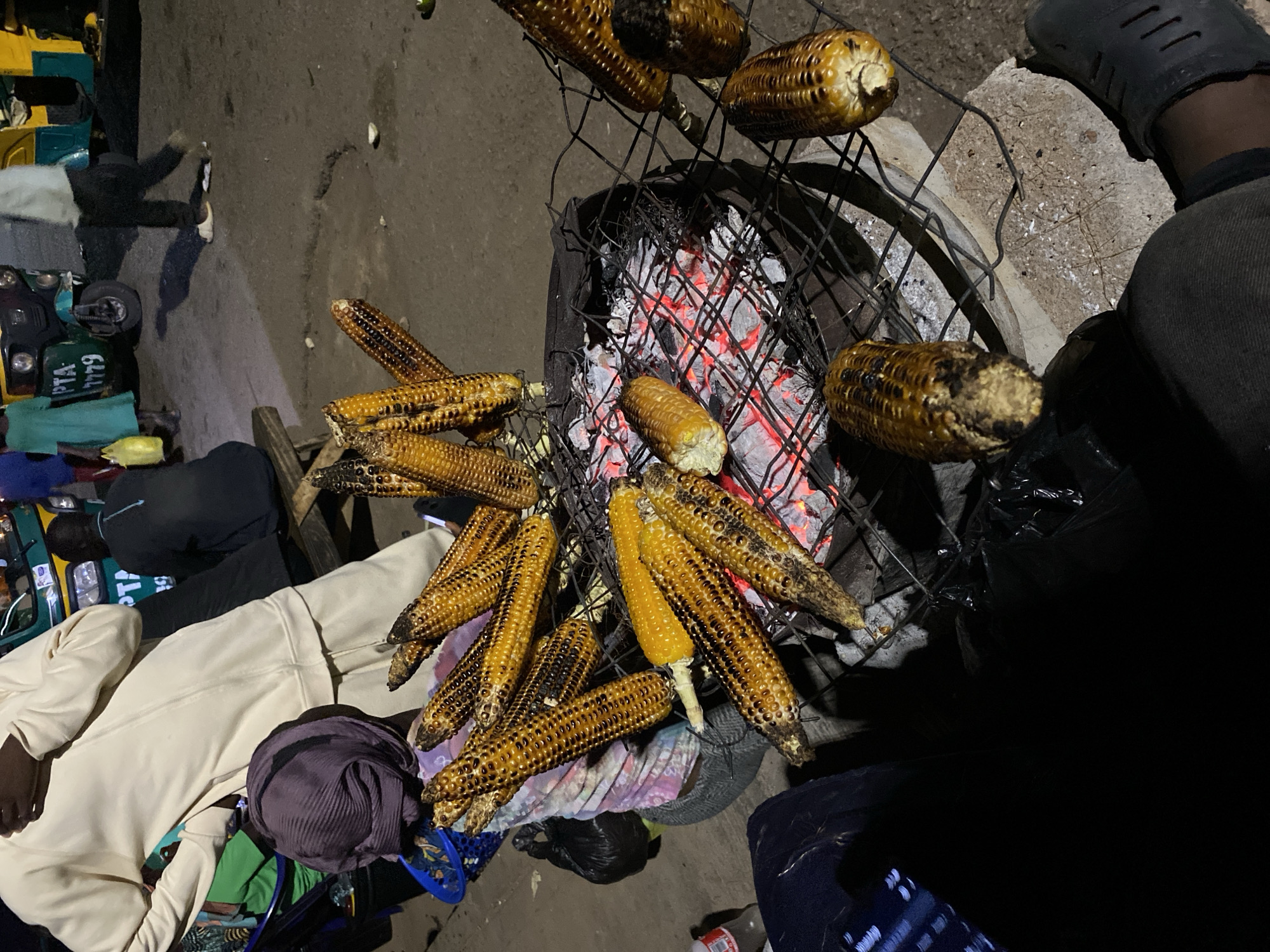The image captures a scene in Bukuru, Nigeria, at night, where grilled corn is being sold. The corn is arranged on a wire grill over a charcoal fire. A person wearing black Crocs and dark trousers sits close to the grill. Next to the grill on the left, a man wearing a beige sweatsuit and a purple headwrap sits on a small stool. Behind him is an auto rickshaw with the number plate PTA 6449. Scattered on the ground are various personal items. The background shows a dim urban setting.