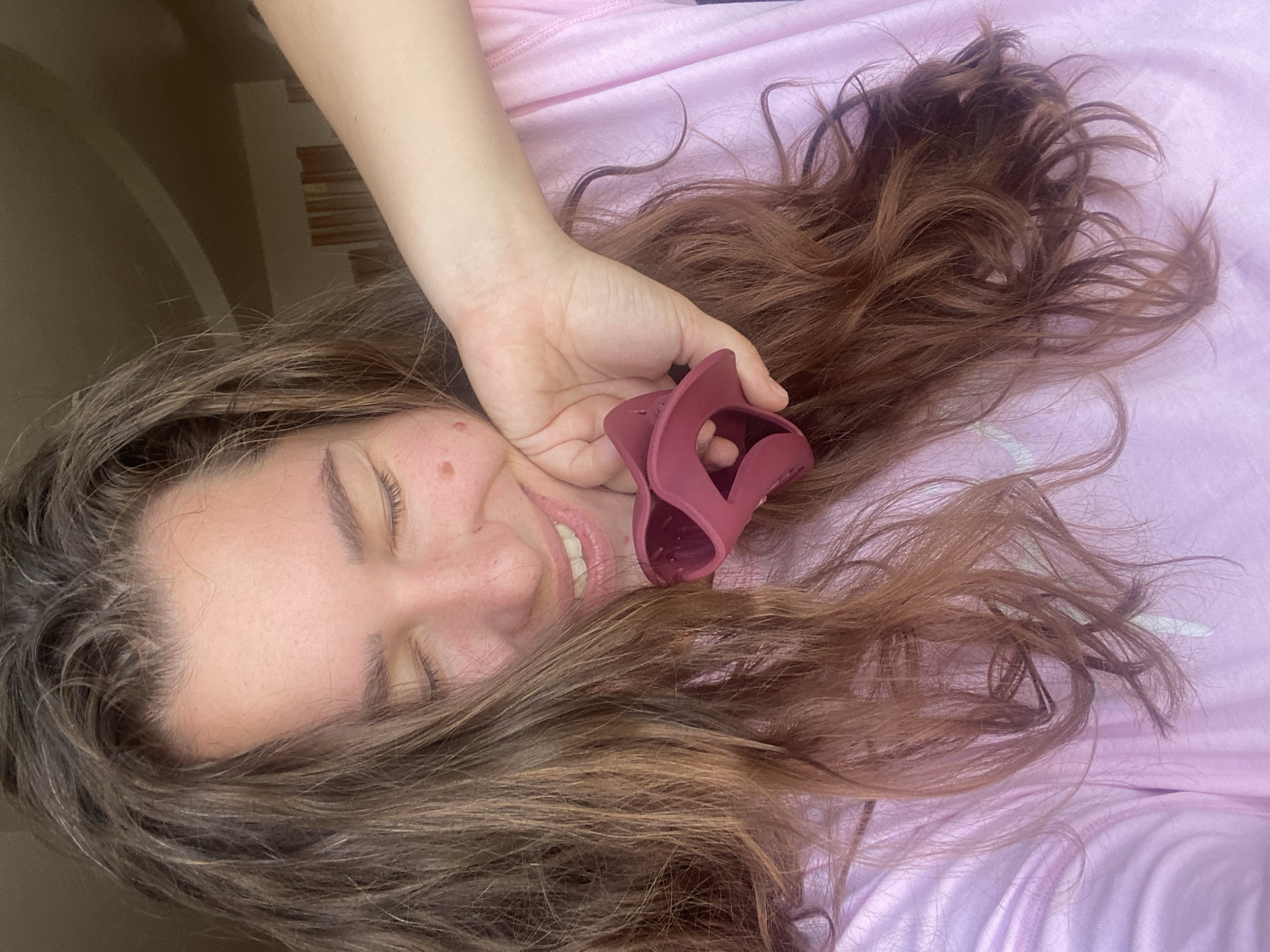 A woman with long, wavy brown hair is holding a maroon hair clip up to her face, smiling with her eyes closed. She is wearing a light pink t-shirt. The background appears to be indoors, with a slanted ceiling and diffused lighting, suggesting a residential setting, possibly in Perm, Russia, although there are no specific visual indicators to confirm the location.