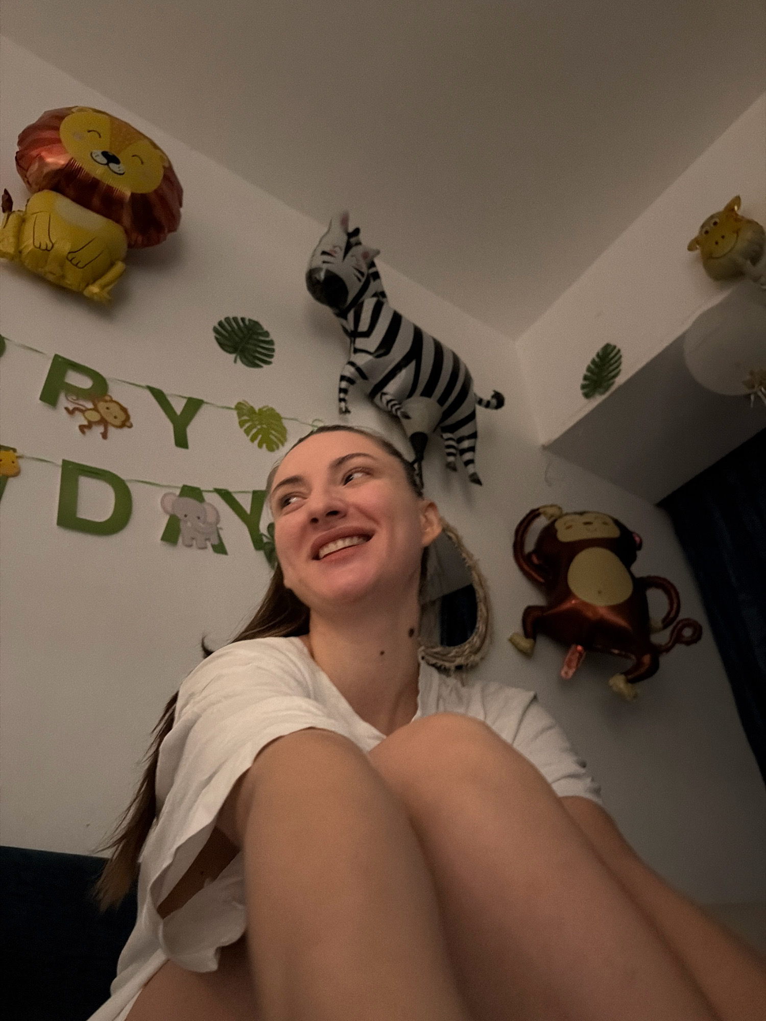 A young woman is sitting in a room decorated with jungle-themed balloons and banners for a birthday celebration. The balloons include a lion, a zebra, and a monkey. There is also a banner that reads "HAPPY BIRTHDAY" with animal cutouts. The woman is smiling and looking upwards. The image appears to have been taken in Sharm El Sheikh, Egypt.