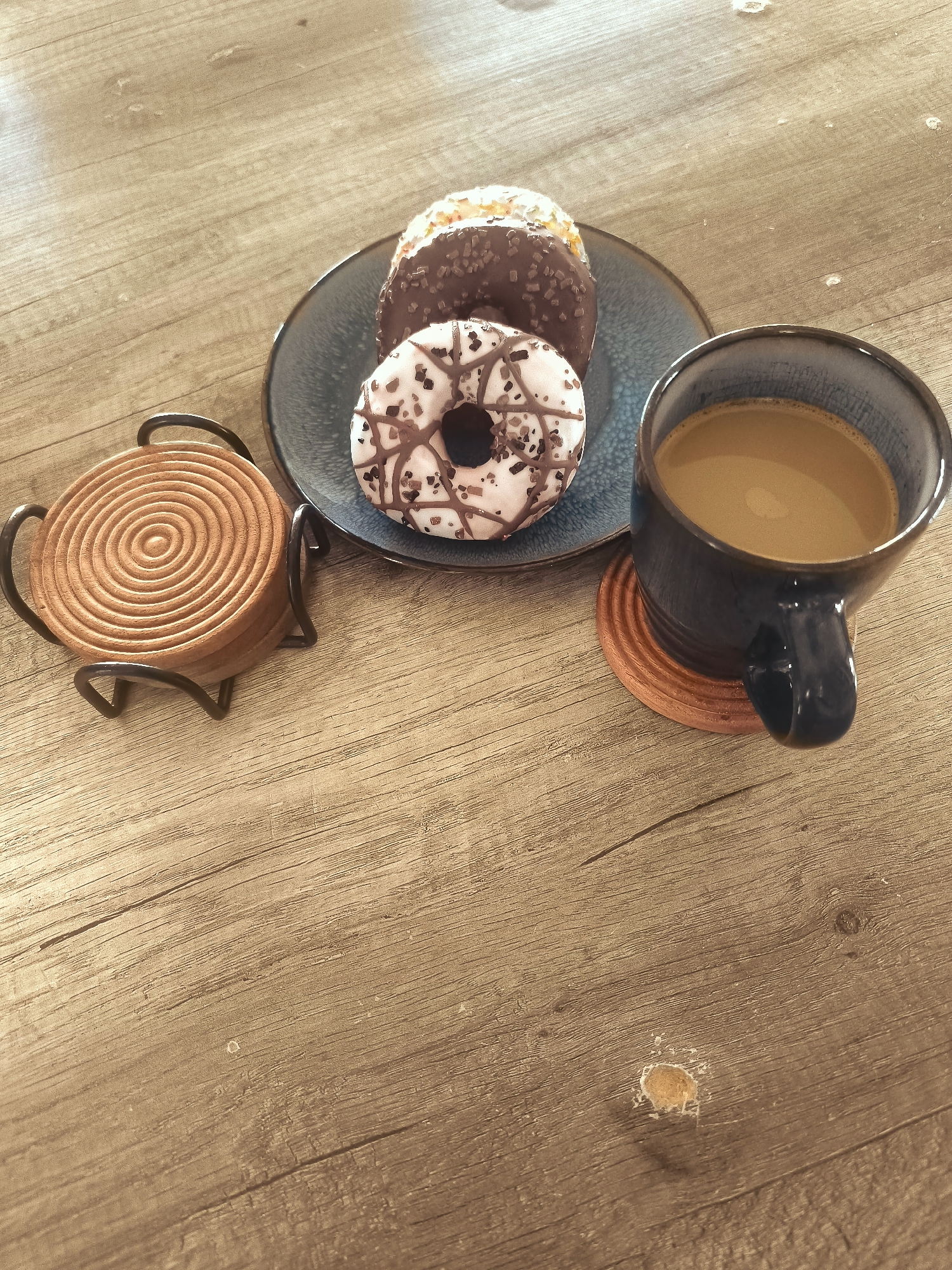 A dark blue ceramic mug filled with coffee is placed on a wooden table. Next to the mug is a blue plate with three donuts. The donuts are of different varieties; one is topped with chocolate and sprinkles, another with white icing and chocolate drizzle, and the third with white icing and brown sprinkles. To the left of the plate is a wooden coaster holder containing stacked wooden coasters. The scene is illuminated by natural light, casting shadows on the wooden surface of the table. The background is out of focus, suggesting an indoor setting. The image captures a moment of leisure, likely a coffee break. The wooden table appears to be a dining table, possibly in a home or a cafe in Almere, Netherlands, given its common architectural style and interior design elements.