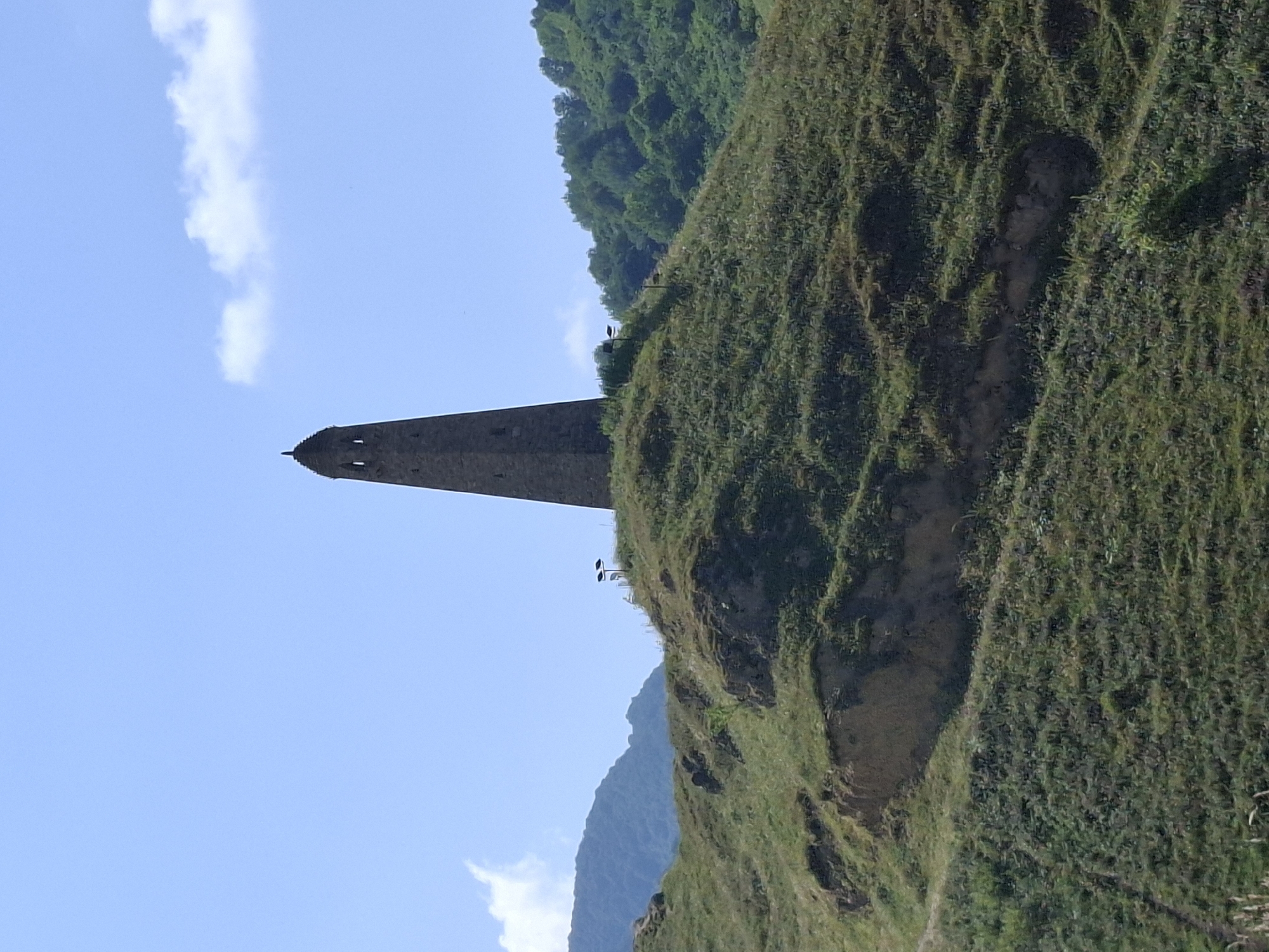 A tall, stone tower stands on a grassy hilltop under a blue sky with scattered clouds. The tower is conical in shape and has two rectangular openings near its top. The hilltop is covered in green grass and some bare patches of earth. In the background, rolling hills covered in trees rise up. Two solar-powered lights are visible on the hilltop near the tower. The scene is serene and appears to be located in a rural area. This tower is identified as the Kura Watchtower, a historical monument located in the village of Kharachoy, Chechnya, Russia.