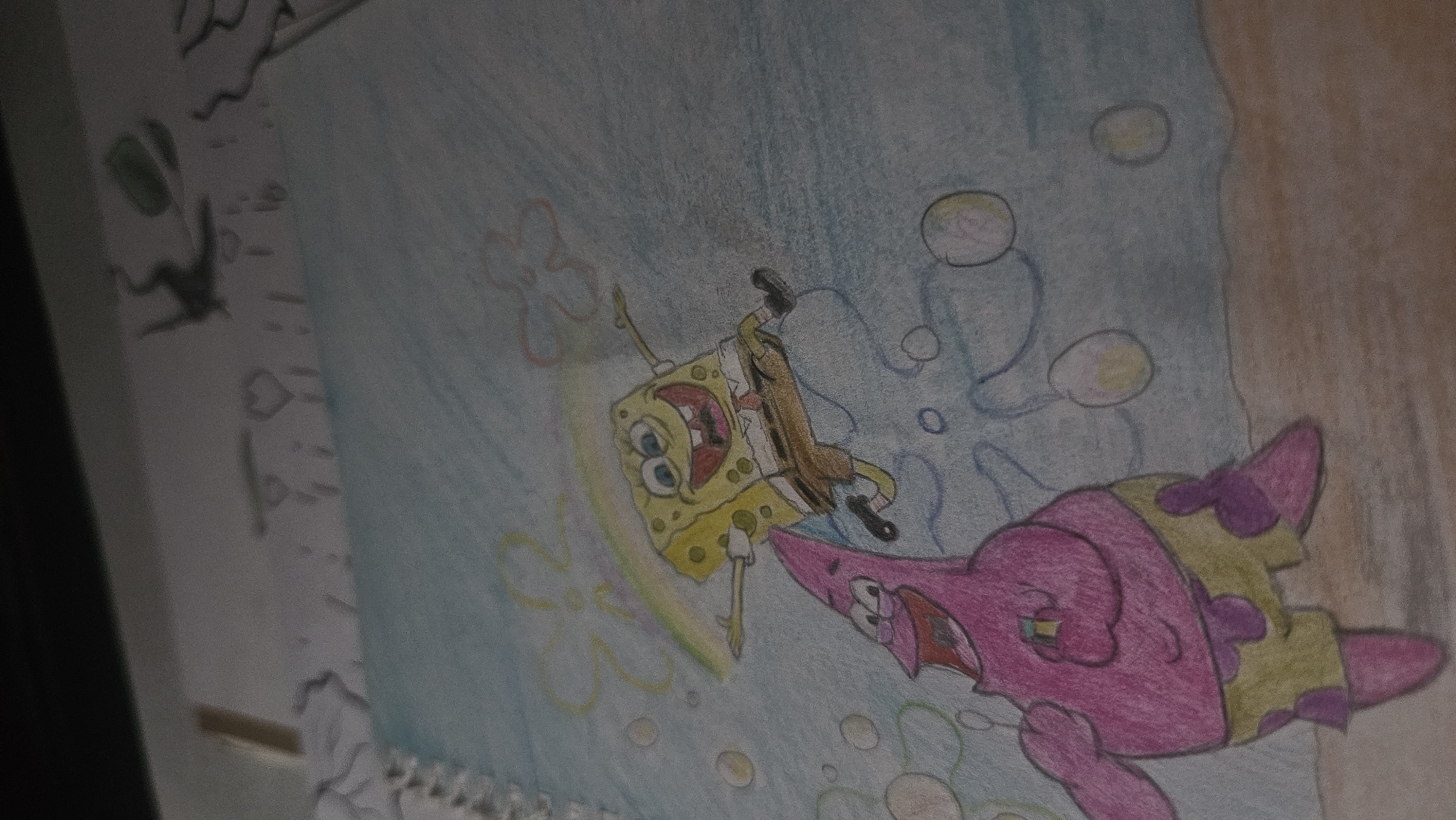 A colored pencil drawing depicts Spongebob Squarepants and Patrick Star in a playful scene. Spongebob, with his characteristic square body, is airborne, arms outstretched and a wide grin on his face. A colorful arc, possibly a rainbow, extends from him. Below him, Patrick, the pink starfish, is also smiling broadly and reaching upwards. Both characters are depicted in a stylized, animated manner. The background is a light blue, suggesting water or sky, with several bubbles scattered around. The bottom of the image shows a sandy brown surface, representing the ground or seabed. The drawing is on a page from a spiral-bound notebook.