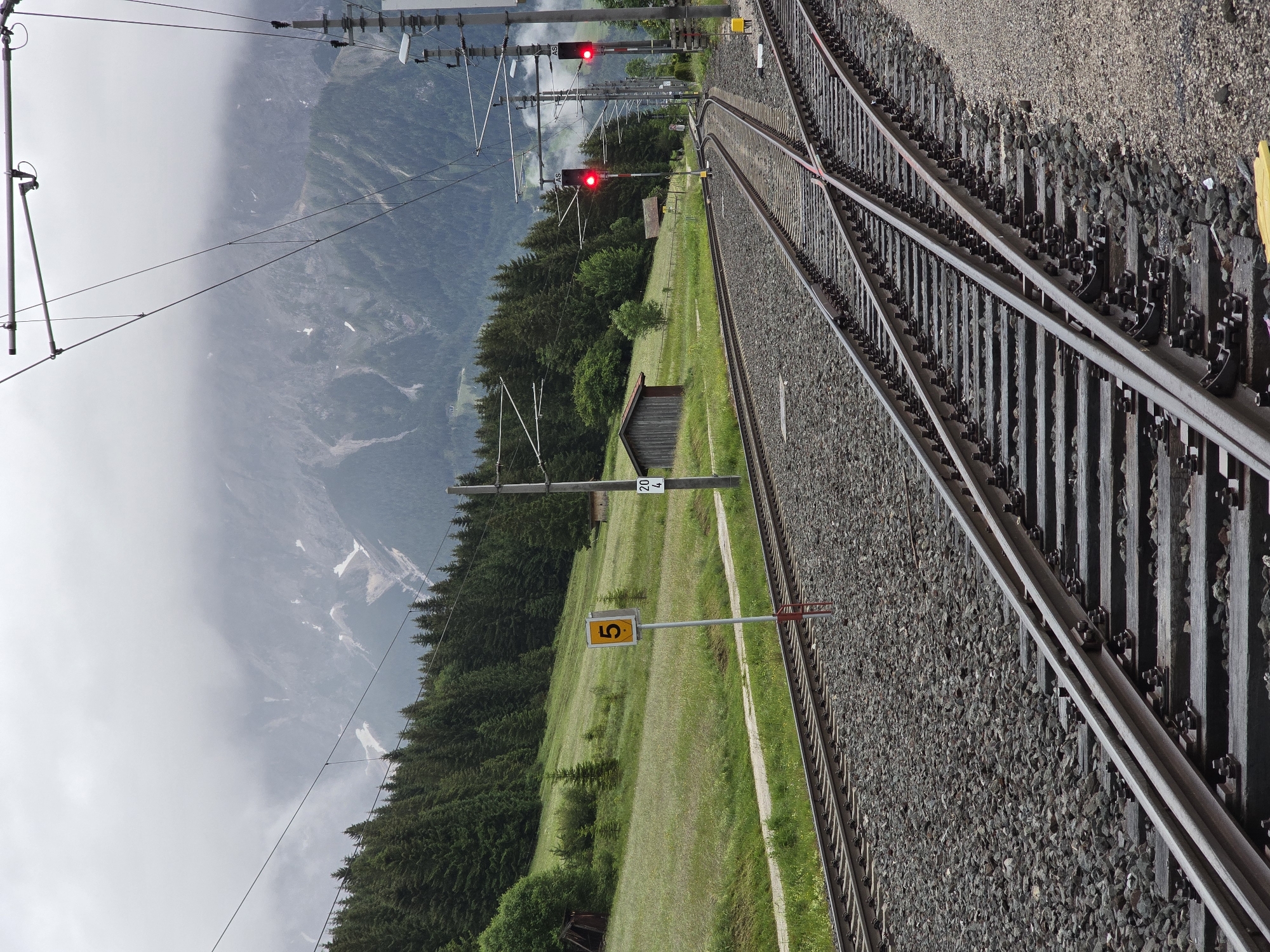 The image depicts a railway line junction in Lermoos, Austria, with the Zugspitze massif visible in the background. The foreground is dominated by railway tracks, gravel ballast, and a complex arrangement of overhead electrical lines. To the left, a single set of tracks runs parallel to the foreground tracks, bordered by a grassy slope dotted with fir trees. A yellow sign with the number "5" is prominently displayed on a pole near the tracks. In the mid-ground, a small wooden shed sits on the grassy slope. Further back, a dense forest of fir trees covers a hillside. In the distance, the imposing Zugspitze massif rises, its peaks partially obscured by clouds. Two red railway signals are visible, indicating that the tracks are currently closed to traffic. The overall scene conveys a sense of a remote, mountainous railway station under a cloudy sky.