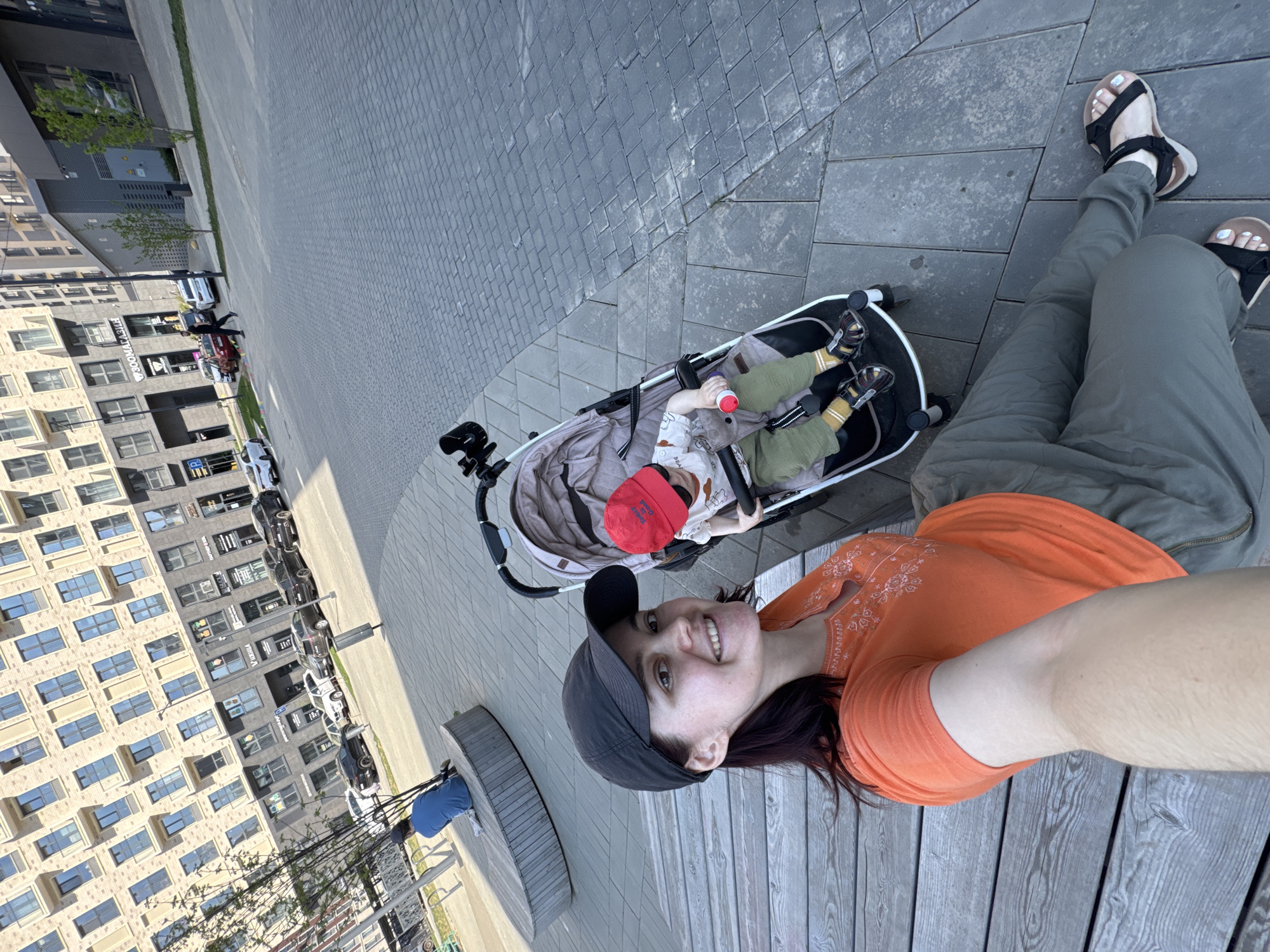 The image shows a woman in an orange shirt and baseball cap taking a selfie with a baby in a stroller. The woman is sitting on a wooden bench. The baby is wearing a red cap and is holding a bottle. The background shows a multi-story building and a paved area. The photograph was taken in Izhevsk, Russia.