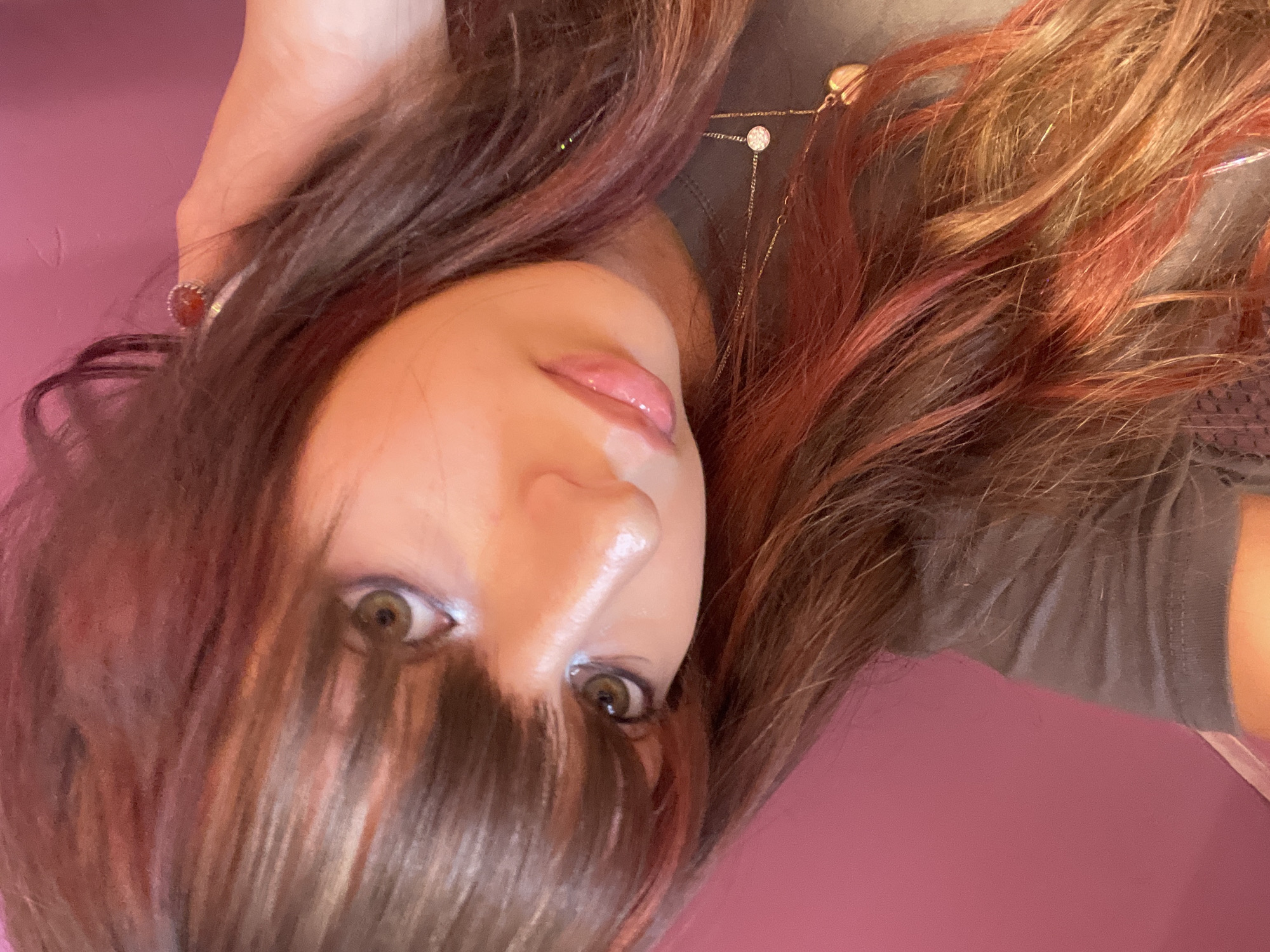 The image is a close-up, tilted selfie of a young woman with long, dark brown hair with red highlights. She is wearing a gray t-shirt and a necklace with a small circular pendant and a heart-shaped pendant. She is also wearing a ring on her index finger. The background is a solid pink wall. The scene appears to be indoors. The woman's face is partially obscured by her hair. She has green eyes and light skin. The image does not contain any information that would allow for the determination of its location in Raanana, Israel.