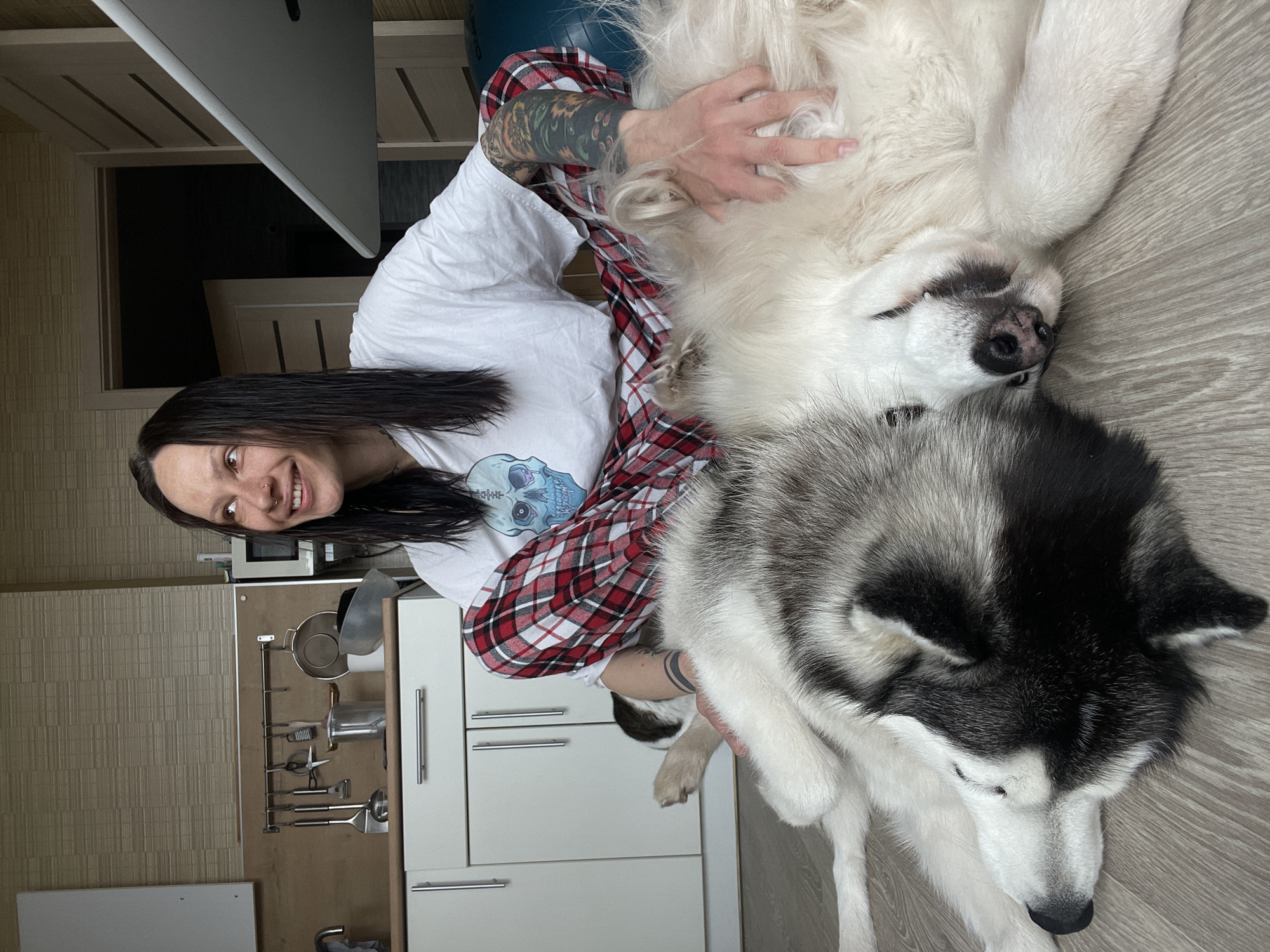 A woman with long dark hair, wearing a white T-shirt with a skull design and a red and black checkered shirt, is smiling at the camera while lying on the floor with two dogs. She is patting a large, fluffy white dog lying on its back. A black and white husky-type dog rests close to her. The scene appears to be indoors, possibly in a kitchen, given the cabinets and utensils visible in the background. The location is Izhevsk, Russia.