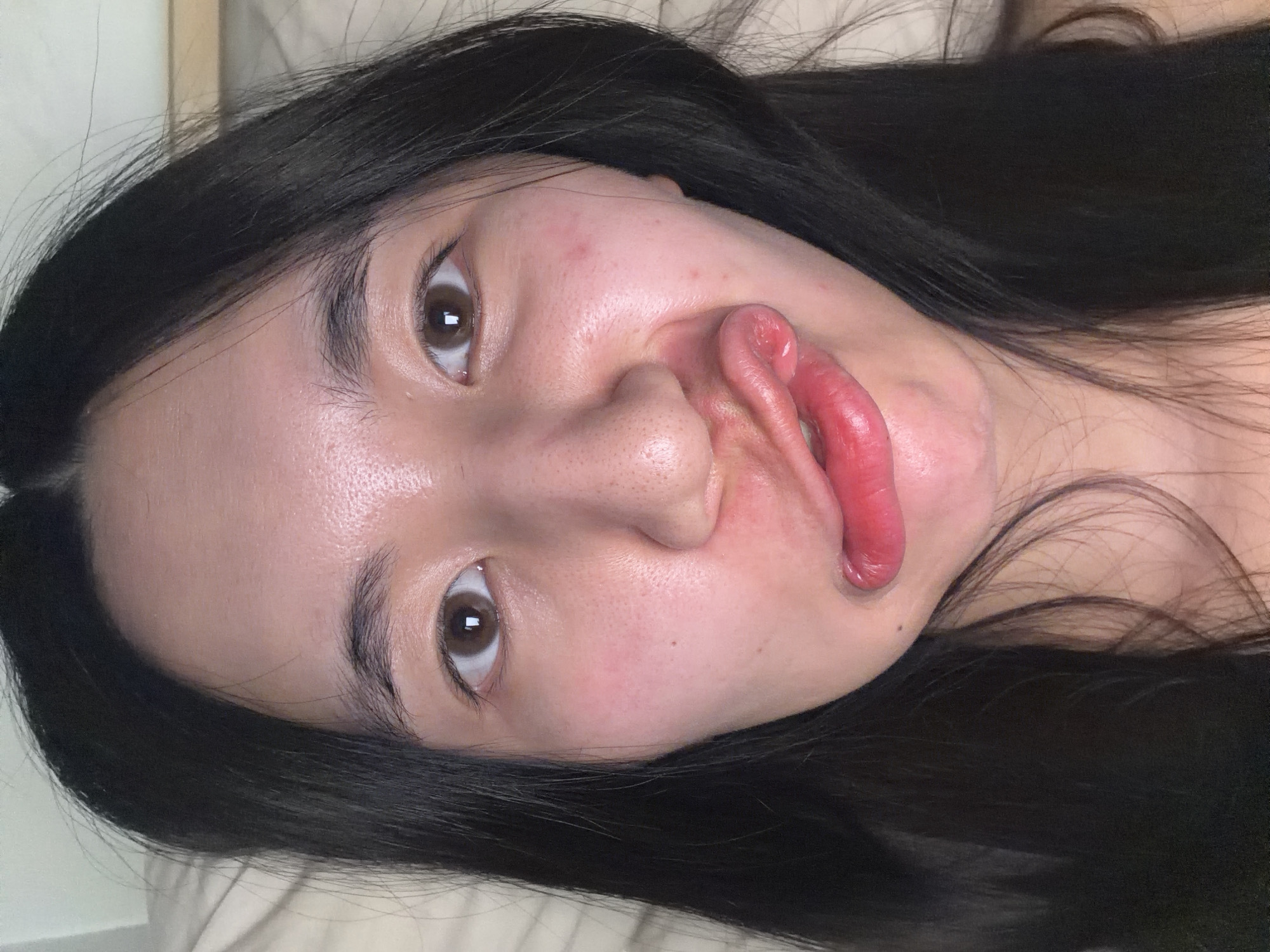 A close-up, angled photograph features a woman with long, dark hair lying on what appears to be a bed covered with a light-colored sheet. Her facial expression is exaggerated, with puckered lips and a slightly distorted nose. The woman has fair skin, brown eyes, and visible pores on her face. There are no visible indicators that the photo was taken in Paris, France, or any specific location. The environment appears to be indoors, possibly a bedroom.