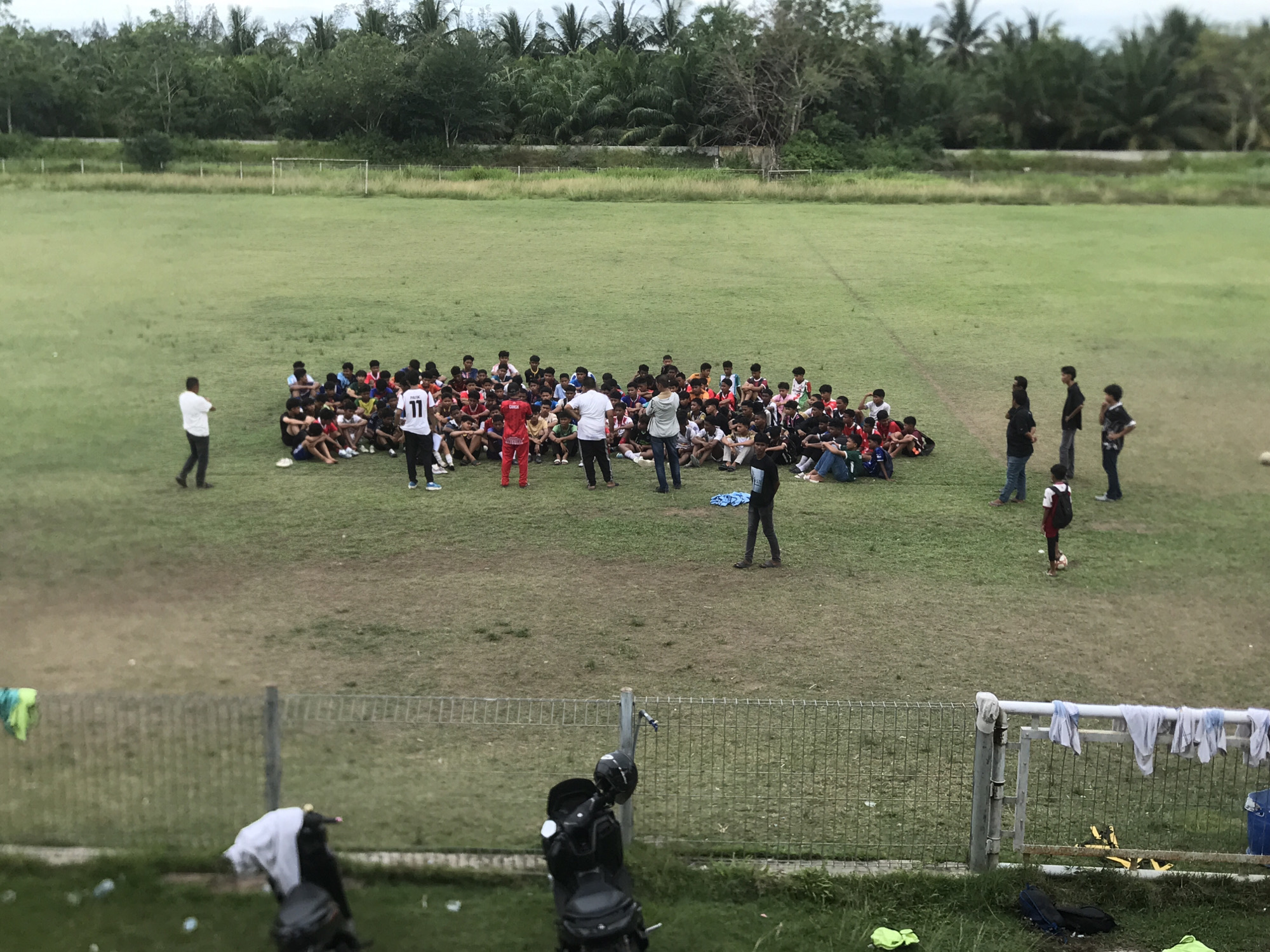 The image shows a large group of people gathered on a grassy field in Tambontunong, Indonesia. The majority of the group is sitting on the ground, arranged in a cluster. Several adults stand among them, appearing to be speaking to the seated individuals. Most of the seated people seem to be young men and teenagers wearing casual clothing. A few individuals stand apart from the main group, with one person dribbling a soccer ball. In the foreground, there is a metal fence, with a motorcycle parked near it. Clothes hang on the fence. The background includes trees and vegetation. A soccer goal is visible in the distance. The sky is overcast.