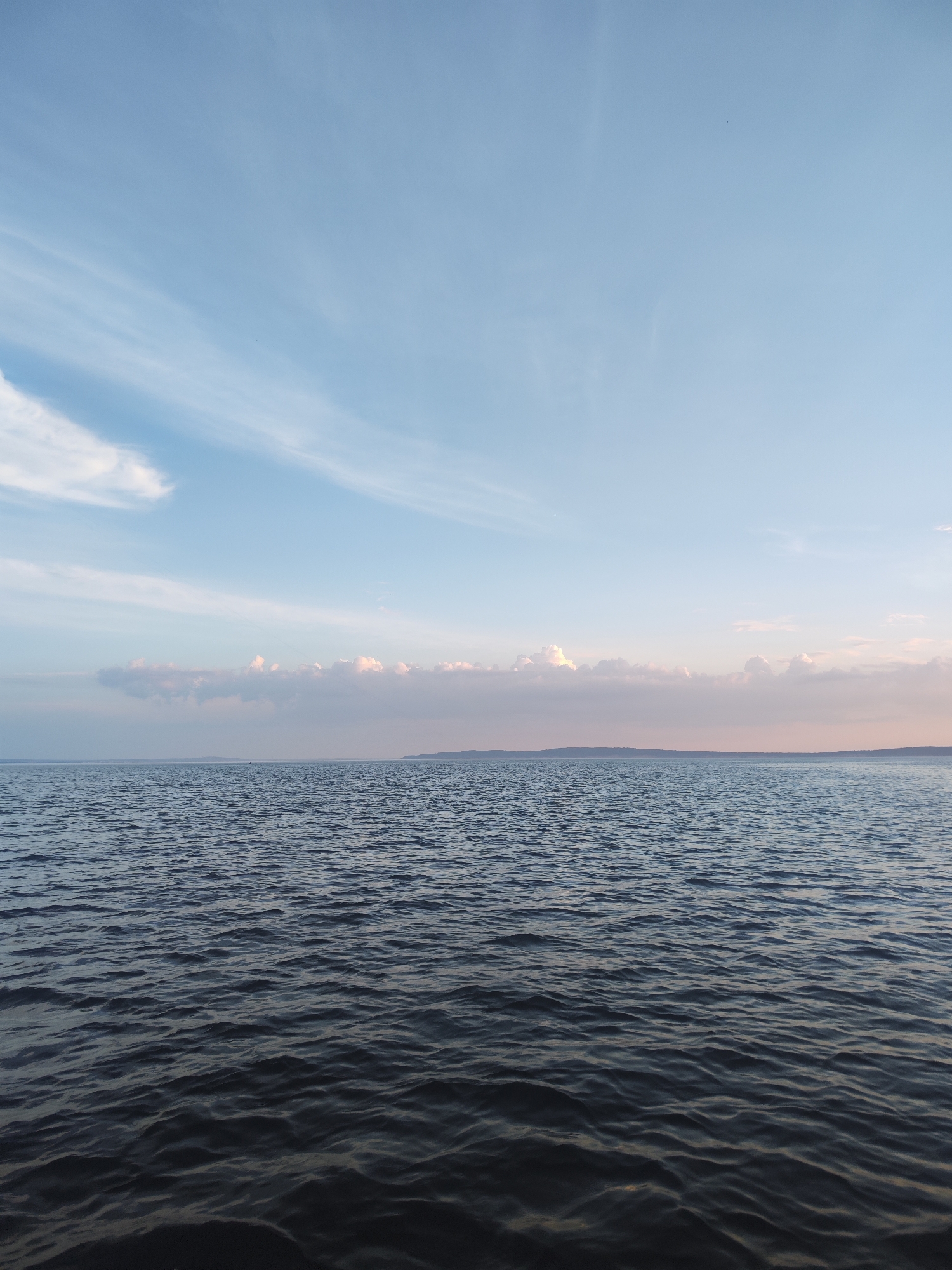 The image depicts a wide expanse of dark, choppy water under a pale blue sky. Wispy white clouds are scattered across the upper portion of the sky, with a band of soft pink and white clouds closer to the horizon. A low, dark landmass is visible in the far distance, forming the horizon line. The water exhibits numerous small waves, suggesting a gentle breeze. The overall scene conveys a sense of open space and tranquility. The location is identified as the city of Galevo, Russia. There are no visible people, specific objects, or discernible actions taking place in the image.