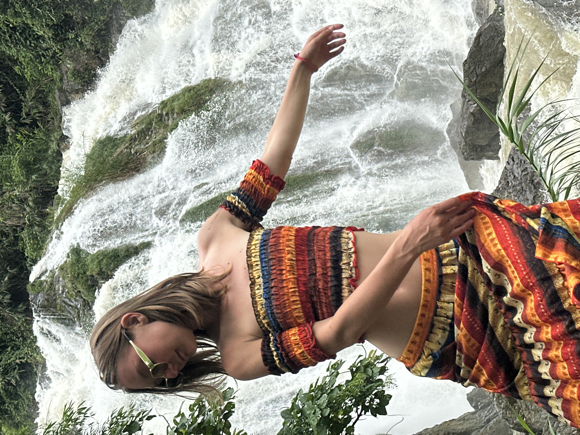 A young Caucasian woman with shoulder-length brown hair, wearing light green sunglasses and a multicolored off-the-shoulder crop top with matching skirt, stands in front of a cascading waterfall in Bản Phúc, Vietnam. Her arms are outstretched, and her body is turned slightly away from the camera. The waterfall features white, frothy water tumbling over moss-covered rocks amidst lush green foliage. The ground in front of the waterfall is rocky, with some vegetation visible. The overall impression is a natural, vibrant outdoor setting.