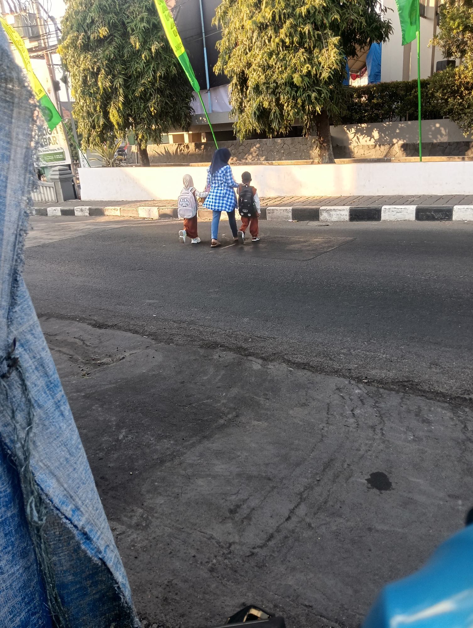 In Winduhaji, Indonesia, at daytime, a person wearing a blue and white checkered shirt and blue pants walks across a street. They are holding the hand of a child wearing a hijab and a light brown outfit, who is carrying a backpack with a cartoon character on it. Behind them, another child, also wearing a light brown outfit and a backpack, walks with the group. The street has a cracked asphalt surface and a paved sidewalk with black and white borders. Tall trees with green leaves line the sidewalk, and a white building with a stone base is visible behind them. Yellow and green flags are attached to poles along the sidewalk. A large piece of blue fabric with torn edges is in the foreground, obscuring part of the scene.