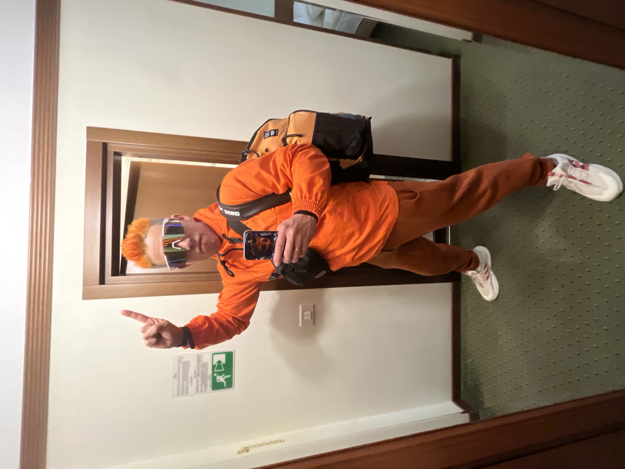 A person wearing an orange tracksuit, futuristic sunglasses, and white sneakers with red stripes is taking a selfie in a mirror. The person has orange hair and is pointing with their left hand. They are wearing a black shoulder bag and carrying an orange and black backpack. The reflection shows a green patterned carpet, a light-colored wall with a light switch, and an emergency exit sign. The location appears to be a hotel room, but there is no definitive evidence to confirm it is in Moscow, Russia.